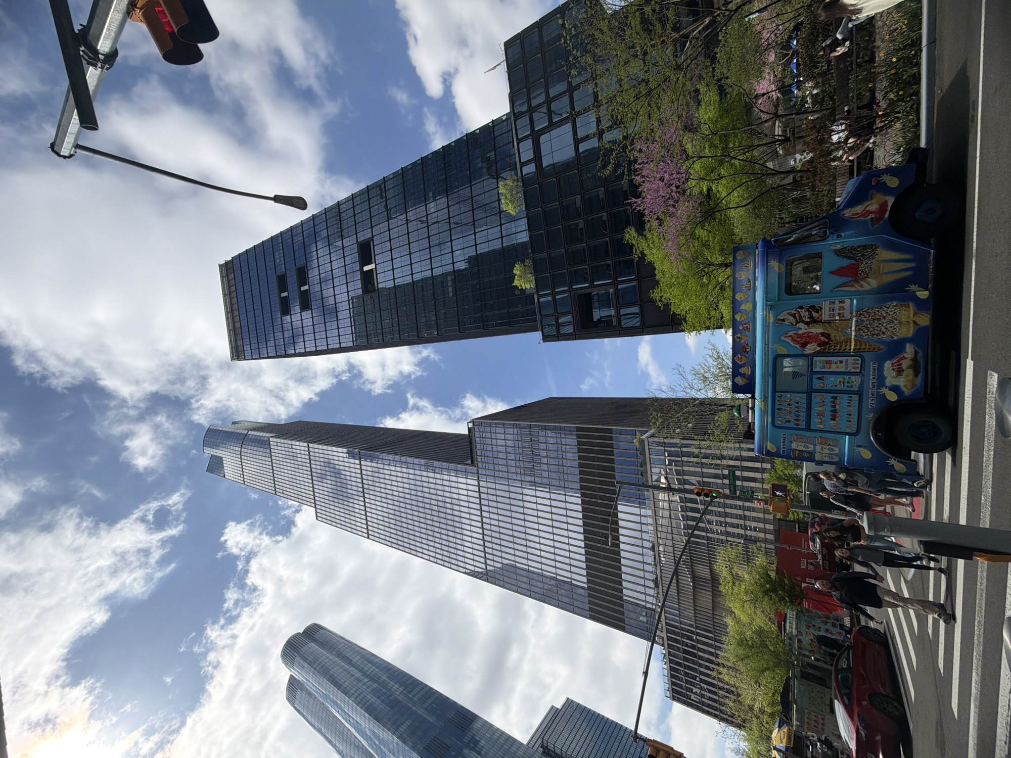 The image captures a vibrant street scene in New York City. Several modern skyscrapers dominate the skyline, with one notable dark-colored building featuring a grid-like facade. Below, a blue ice cream truck stands parked alongside the curb, its exterior adorned with colorful images of frozen treats. Pedestrians can be seen walking across a crosswalk, suggesting a bustling urban environment. A traffic light is also visible. The presence of flowering trees with pink blossoms adds a touch of springtime color to the scene. The composition of the image creates a sense of looking up towards the tall buildings, emphasizing their imposing height.