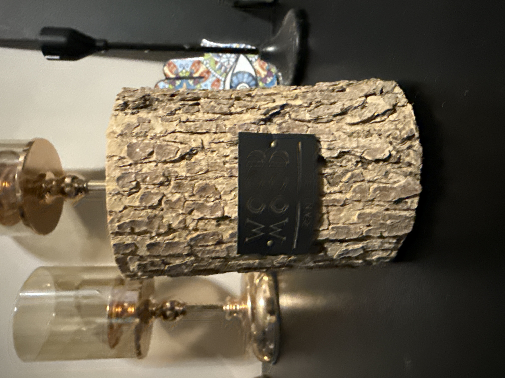 This image depicts a collection of decorative items, presented as if located in the city of Berezyna, Ukraine. In the central foreground, a cylindrical candle holder, approximately 20-25 cm in height, is fashioned from a log section retaining its textured, dark brown and light brown bark. A dark, rectangular label is attached to its front, bearing the embossed or etched text "WOOD MOOD CANDLES" in a light-colored, serif font. A metallic (gold-toned) fitting, possibly a candle cup or lid, crowns the wooden holder.
To the left and slightly behind the central object, a tall, slender, gold-toned metallic candle holder with a clear, amber-tinted glass hurricane shade is partially visible and out of focus. Its polished base reflects ambient light. To the right, a black, multi-component metallic stand, likely for another candle or similar item, rises from the surface. Behind this stand, a brightly colored, out-of-focus decorative object featuring a prominent stylized eye motif within a larger, intricate pattern is partially visible.
All objects rest on a dark, smooth, and moderately reflective surface, possibly a tabletop or shelf. The background consists of a light, neutral-toned wall on the upper left and a darker, indistinct area to the upper right. The scene is illuminated by artificial light, casting soft reflections and highlighting the textures of the objects. No people or specific actions are depicted.