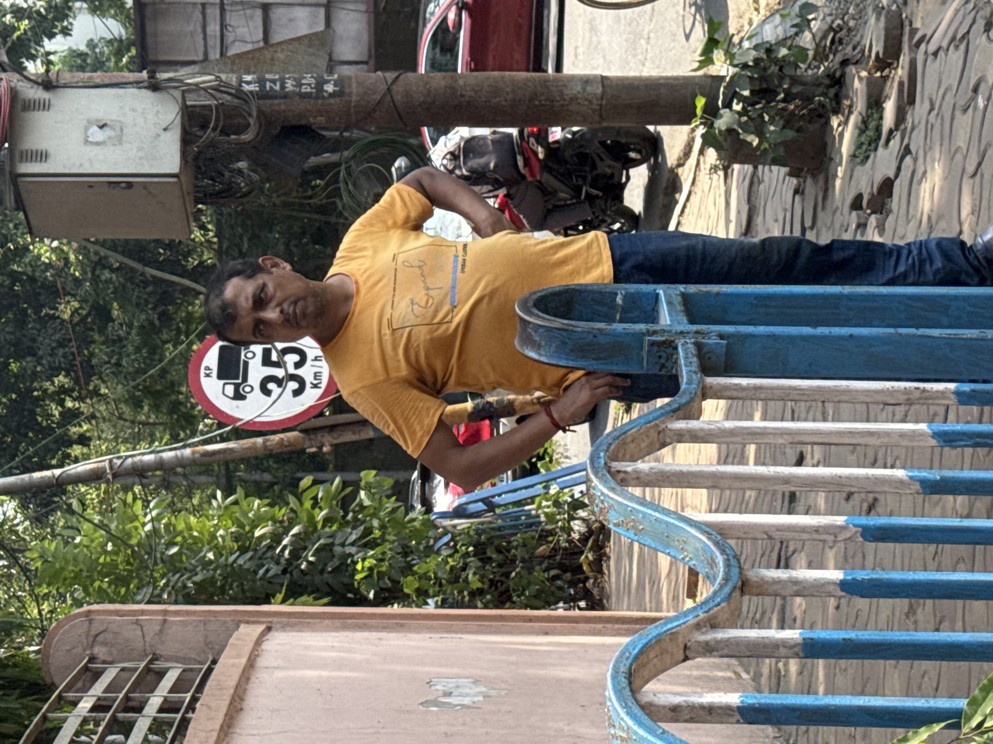 The image depicts a man standing next to a blue and white painted metal railing in what appears to be a street scene in Kolkata, India. The man is wearing a yellow t-shirt and blue jeans, and he is leaning against the railing. A speed limit sign indicating 35 km/h is visible in the background. Behind the man is a motorcycle and a red vehicle. The area includes trees and a building with a window. The location seems to be a roadside or park area, given the presence of foliage and the building. Overhead, there are utility poles and cables.