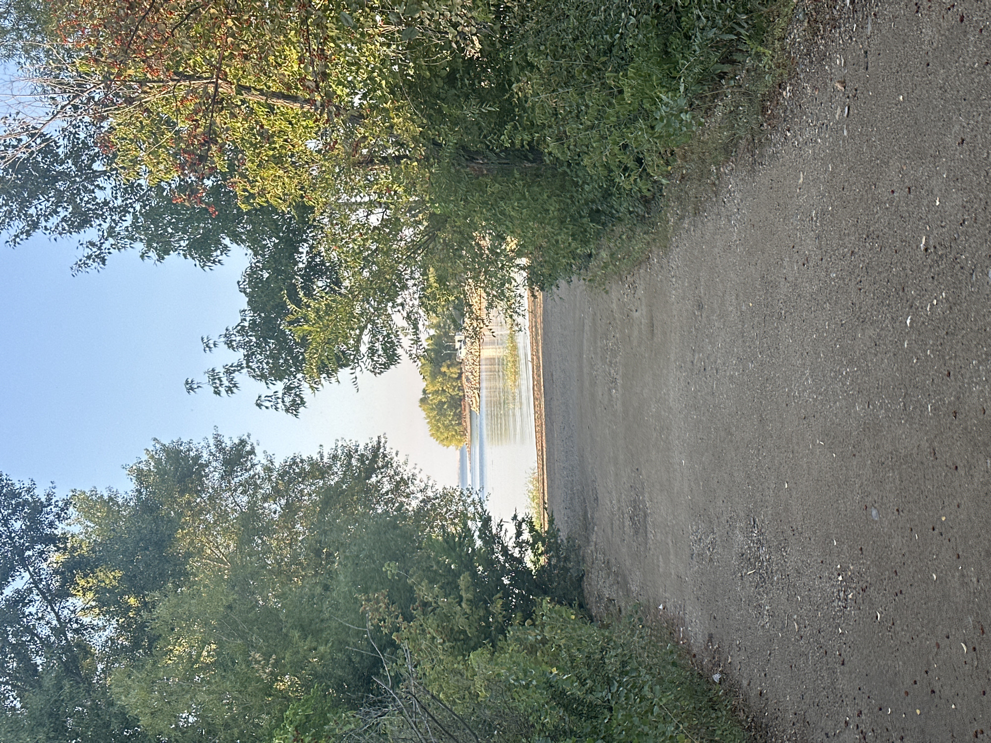 This image depicts a pathway leading towards a body of water, possibly a reservoir or lake, framed by lush green trees on either side. The pathway is unpaved, appearing to be made of gravel and dirt, with scattered leaves and small debris. The water is calm, reflecting the clear blue sky above. In the distance, across the water, there are trees lining the shore of a landmass. The overall scene suggests an outdoor, natural setting, potentially a park or a recreational area. The location in the city of Energetik, Russia, is not discernible from the visual information in the image. There are no people or specific structures that would indicate this specific locale. The lighting suggests it is daytime, possibly late afternoon due to the quality of light filtering through the trees.