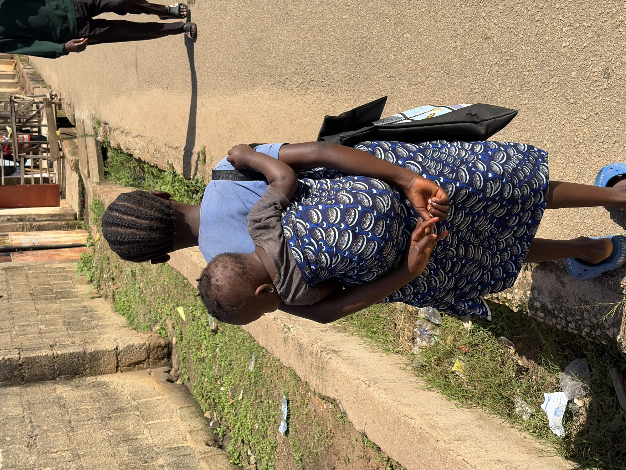 In Jos, Nigeria, a young girl, likely a pre-teen or early teen, carries a baby on her back, secured with a patterned cloth wrap. The baby is swaddled and appears to be asleep or resting. The girl is wearing a blue t-shirt and a blue and white patterned dress or skirt. She has her hands clasped behind her back. Her footwear consists of blue slip-on sandals.
The scene unfolds on an outdoor path or unpaved road. To the left, a rough, textured concrete wall runs along the side of the path, with overgrown grass and scattered debris at its base, including plastic wrappers and bottles. Beyond the wall, a ditch or drainage area is visible, with some vegetation and a narrow concrete bridge crossing it. In the background, structures with wooden walkways and corrugated metal roofs suggest a residential or market area.
To the right of the girl, the path is a dusty, light brown color, with a few scattered pebbles. Further down the path, a person wearing a dark green long-sleeved shirt and dark pants, with black sandals, is walking away from the camera. The sunlight casts distinct shadows on the ground, indicating a bright, sunny day. The overall impression is a snapshot of daily life in a bustling, possibly developing, urban environment in Nigeria.