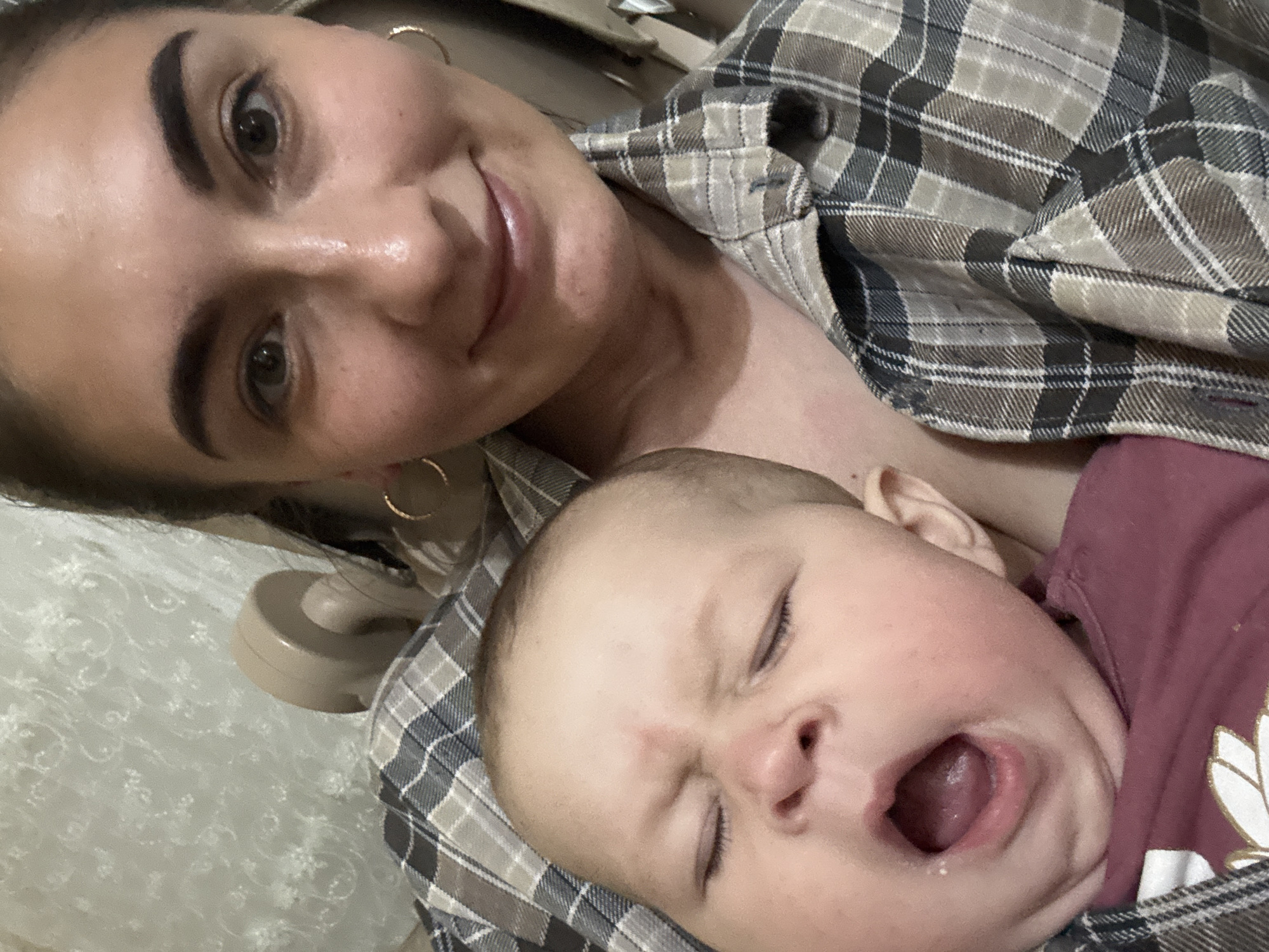 A selfie shows a woman and a baby. The woman, fair-skinned, has dark eyebrows, dark hair pulled back, and is wearing hoop earrings and a gray and brown plaid button-down shirt. She is smiling slightly. The baby is fair-skinned and appears to be yawning or crying with their eyes closed and mouth open. The baby is wearing a maroon shirt. A beige object, possibly a phone, is partially visible in the background. The location is not discernible from the image. There is no information to suggest the location is in Sochi, Russia.