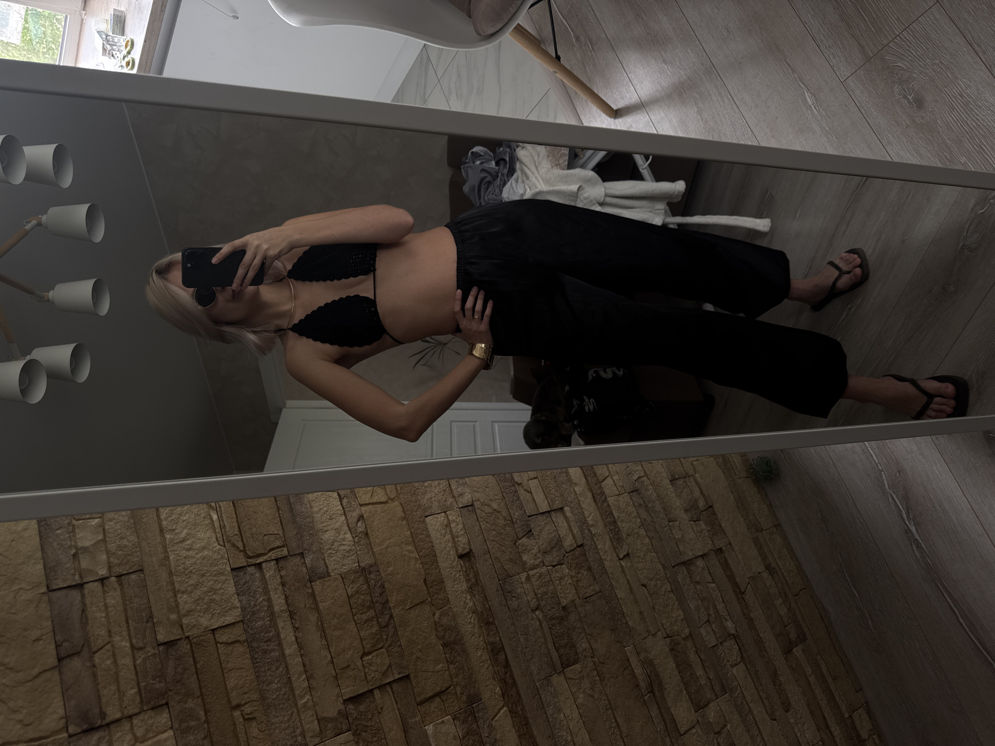 A mirror selfie shows a woman with short, blond hair wearing a black bra top and loose black pants. She holds a black smartphone, obscuring her face, and wears sunglasses and a gold bracelet. Her body is angled towards the viewer, with one hand on her hip. She wears black flip-flops. The reflection shows a room with a hardwood floor and a partial view of a white chair, a clothing pile, and a doorway. The wall adjacent to the mirror is covered in brick-like tiles. The lighting fixture is visible on the left. The location is unspecified and cannot be determined from the image, nor can we confirm that the image was taken in Saratov, Russia.