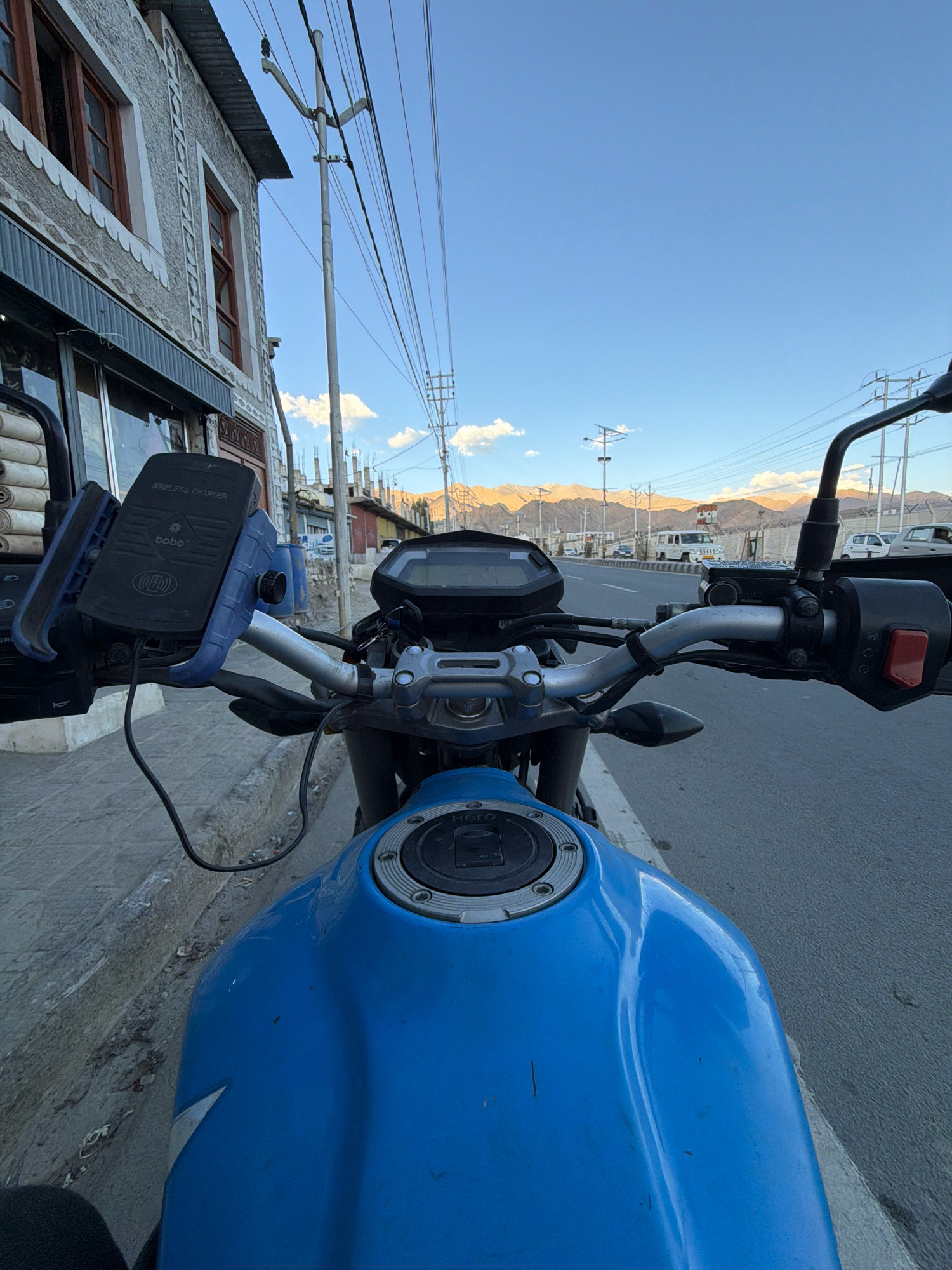 The image shows a first-person perspective from a motorcycle parked on the side of a street in Leh, India. The motorcycle is bright blue and has a wireless charger mounted on the handlebars. The street appears to be paved, with a sidewalk made of concrete and gravel to the left. In the background, there are buildings with traditional Tibetan architectural elements, including wooden window frames and decorative patterns on the walls. Utility poles with numerous electrical wires are visible, a common sight in Indian cities. Further in the distance, mountains form a backdrop against a clear blue sky with a few scattered clouds. A few vehicles, including a white SUV, are present on the road. The time of day appears to be late afternoon or early evening, given the warm light illuminating the mountains. The scene suggests a typical urban environment in Leh, likely a residential or commercial area.