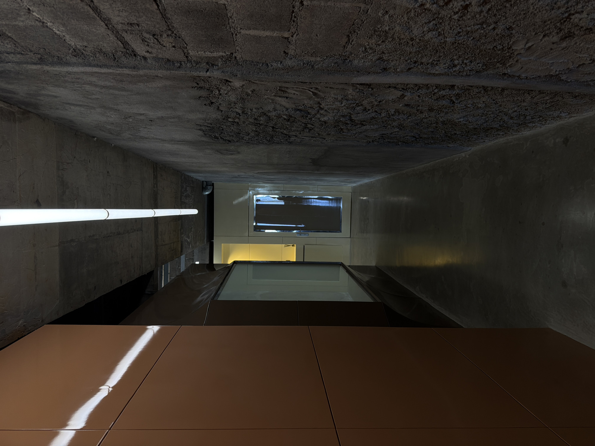 The image presents a view from below, looking upwards into a narrow architectural space, possibly a light well or courtyard. The bottom foreground is dominated by brown, square tiles with a glossy finish, reflecting a distinct line of light. Above the tiles, a dark gray or black angled structure leads towards a smaller, rectangular opening revealing daylight and the facade of a building beyond. Within this opening, a window with closed blinds can be seen. The walls enclosing this space are characterized by rough, concrete surfaces with varying textures. A horizontal fluorescent light fixture is visible on the left wall. The top portion of the image shows a raw concrete ceiling with a coarse texture. There are no people visible. The location, North Kuta, Indonesia, is not directly evident from the visual information provided.