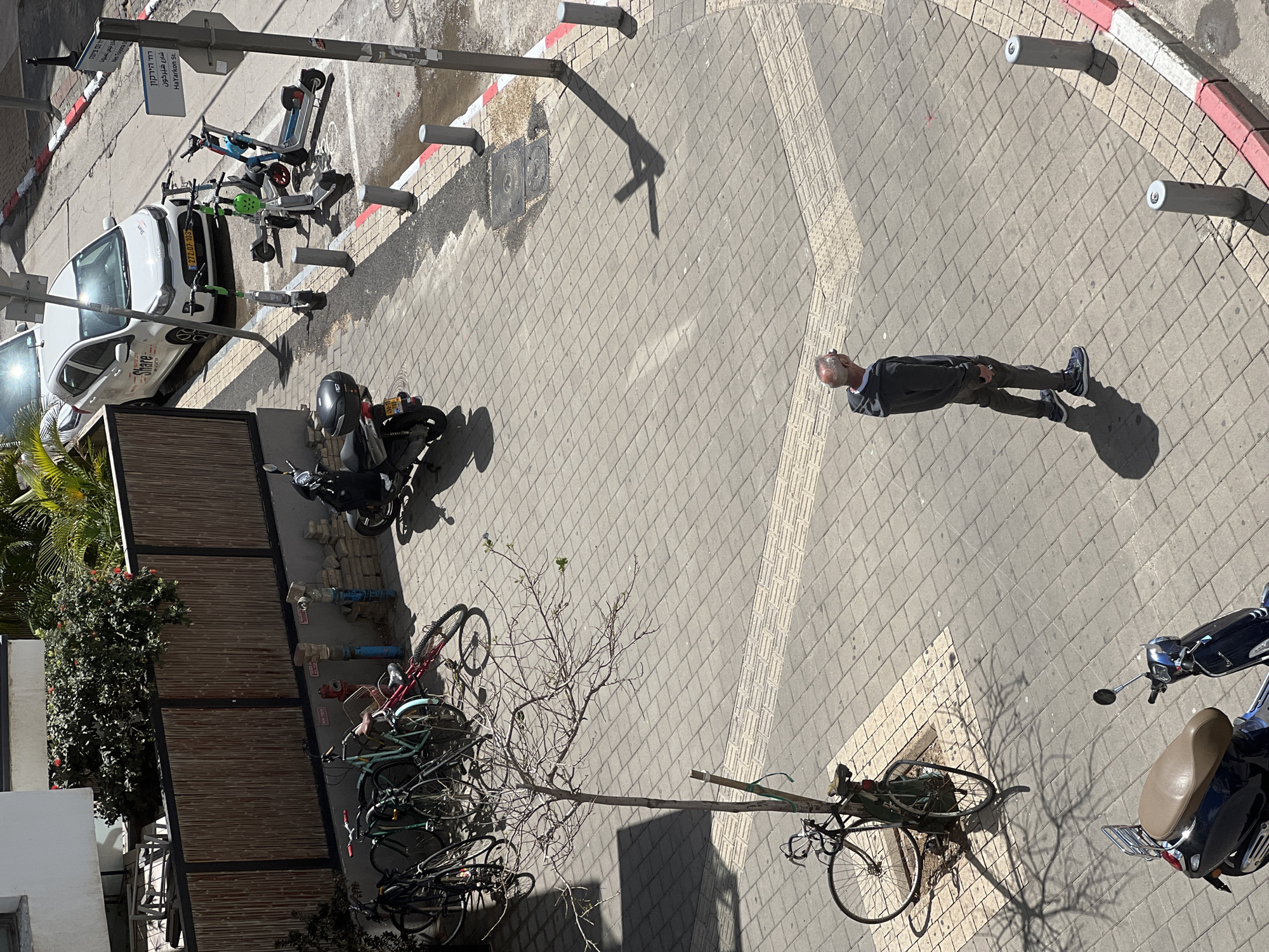 The image, taken from a high angle, shows an urban street scene in Tel Aviv, Israel. At the top left, there's a parked white car with "Share" displayed on its side and a license plate number "272:07-103". Next to it, there is a cluster of electric scooters.
Parallel to the car is a sidewalk area leading to the intersection that curves at the top right. The intersection has a red and white curb and vertical grey posts. A man stands on the sidewalk near the intersection. He is wearing dark clothing, trousers, and blue sneakers.
A motorcycle is parked near a wooden fence. Beside the fence, a stack of bicycles are visible, leaning against each other and a bare-branched tree. At the bottom right, another motorcycle is parked. Street signs are also visible with Hebrew and English writing, including the street name "HaYarkon St.".