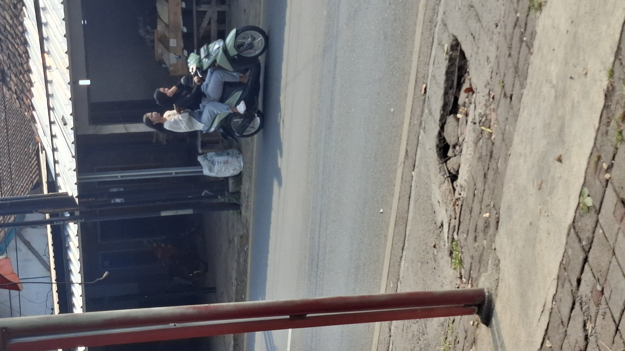 Two young women are riding a light green scooter on a street in Cilaku, Indonesia. The woman in front is wearing a black headscarf and a dark top. The woman behind her is wearing a white cardigan and light blue pants. They are both looking to the right. The scooter is moving on a paved road. In the background, there are buildings with tiled roofs and some shops. A large pothole is visible on the side of the road, with exposed bricks and metal rebar. A red pole is in the foreground on the left side of the image.
