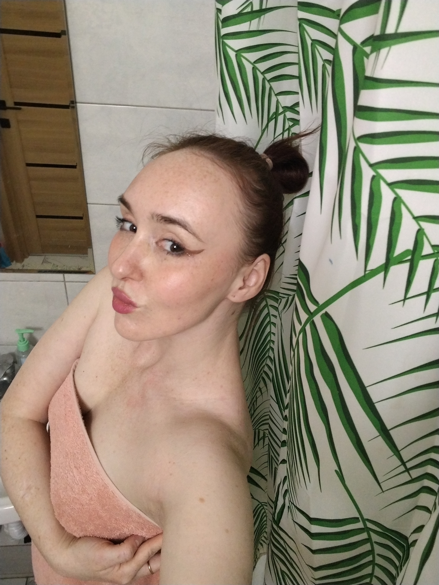 A young woman with light skin and numerous freckles on her face, shoulders, and chest is pictured in what appears to be a bathroom. Her dark hair is pulled back into a messy bun. She has prominent winged eyeliner and pink lipstick, and her lips are pursed in a playful "kiss" expression as she looks towards the camera. She is wrapped in a light peach-colored towel that covers her torso. On her left hand's ring finger, a simple golden ring is visible.
The background consists of light-colored square tiles on the left, next to what appears to be a wooden door or cabinet with horizontal black accents, possibly reflected in a mirror. A light green pump bottle is visible on a white surface near the bottom left corner, suggesting a sink or shelf. To the right, a white shower curtain with a vibrant green palm leaf pattern hangs, partially obscuring the background. The overall setting is an indoor bathroom, and the woman appears to be taking a selfie, likely after a shower or during a beauty routine. No specific time of day or external location cues are discernible from the image itself, beyond the general indoor setting. There is no visible text.