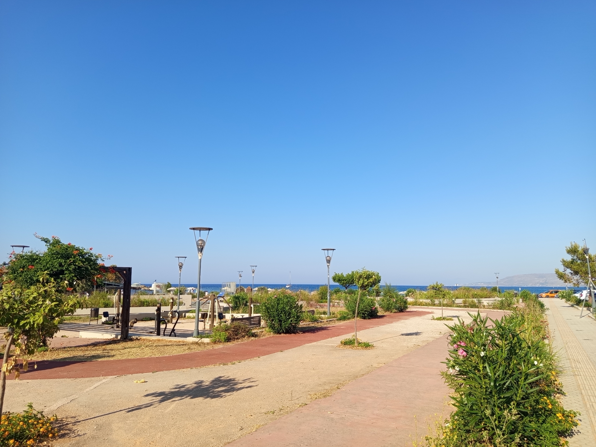 This image depicts a section of the waterfront promenade in Georgioupoli, Greece, on a clear, sunny day. The foreground features a paved pathway with distinct red and beige sections, lined with low-lying green foliage and small trees. Several lampposts with modern designs are strategically placed along the path.
Further back, an open-air exercise area is visible, equipped with various gym machines and benches. A lush green tree with orange flowers stands to the left, adding a splash of color to the scene. Beyond the exercise area, the pathway curves towards the sea.
In the background, the deep blue Aegean Sea stretches out, with a sailboat visible on the horizon. The coastline is dotted with sparse vegetation and appears to be a popular area for recreation, with a few people strolling or relaxing near the water's edge. The overall atmosphere is serene and invites outdoor activity.