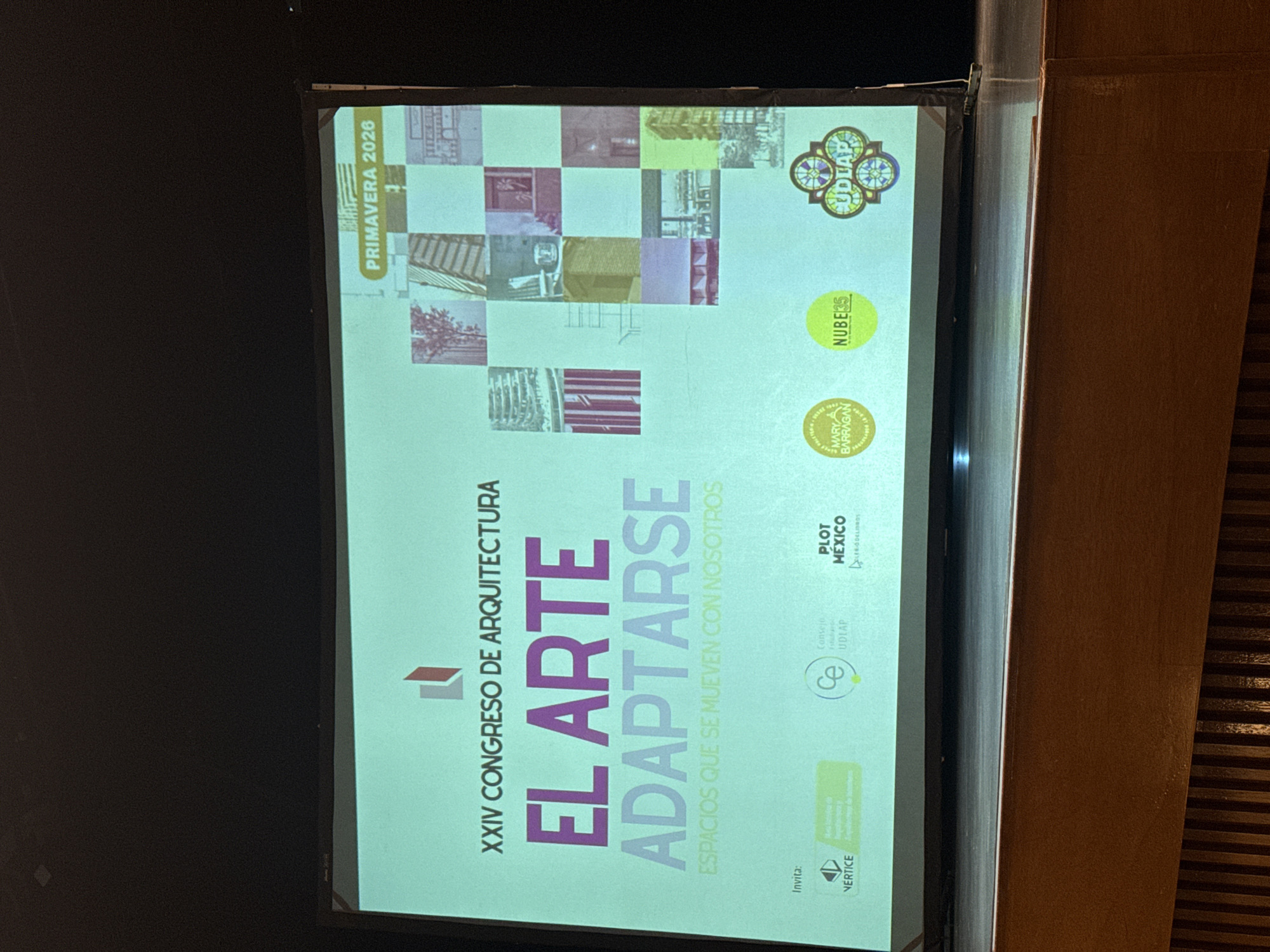 A photograph shows a screen displaying the poster for the XXIV Congress of Architecture, titled "El Arte Adaptarse" (Art Adapt). The text reads, "Espacios que se mueven con nosotros" (Spaces that move with us). The poster also notes it's Primavera (Spring) 2026. Sponsor logos include Nube 35, Plot Mexico, Vertice, UDLA (Universidad de las Américas Puebla).
UDLA is located in San Andrés Cholula, Mexico.