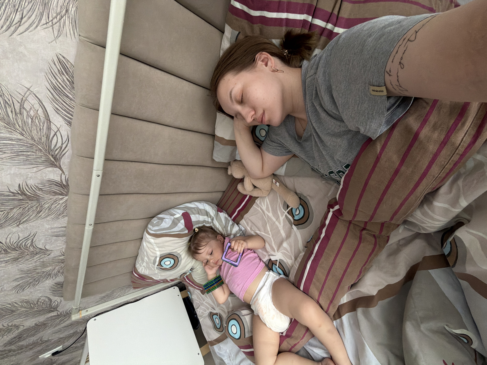 The image depicts a woman and a baby sleeping on a bed. The woman, with brown hair tied back in a ponytail and wearing a gray T-shirt, is lying on her side with her eyes closed. A stuffed animal is nearby. Next to her, a baby wearing a pink top and diaper is also asleep, clutching a purple object. The bed is covered in patterned sheets and pillows. A white table stands next to the bed. The location is Reshetnikovo, Russia.