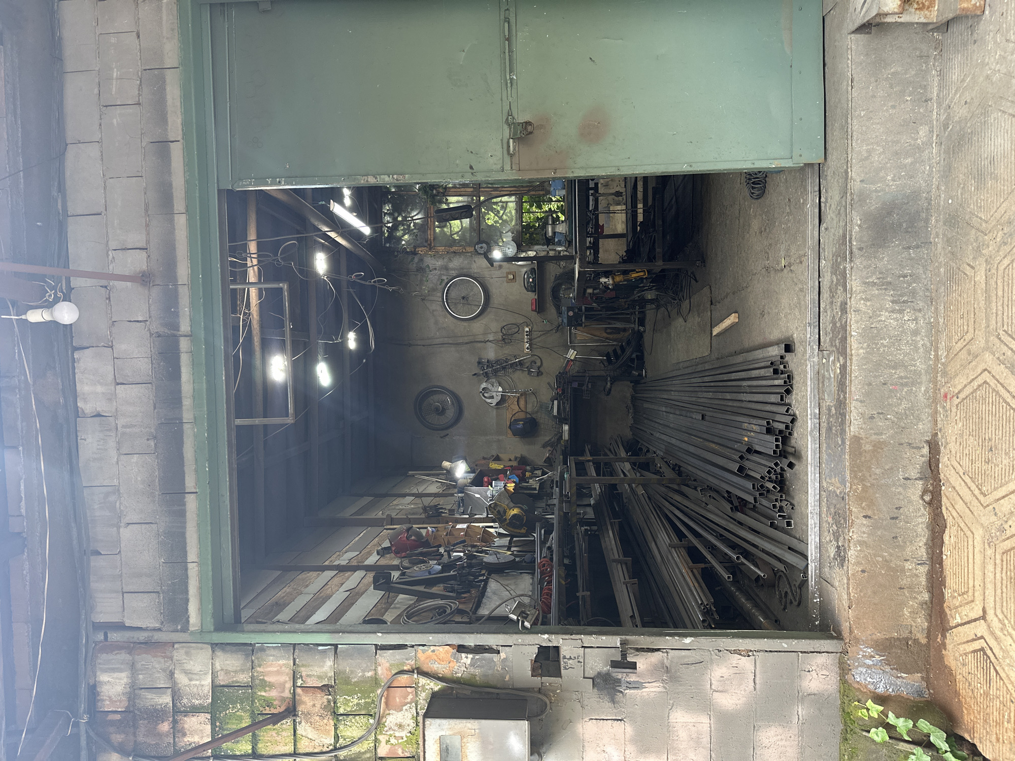 The image depicts a workshop, possibly in Tbilisi, Georgia. The workshop is accessed through a large, partially open, green metal door. Inside, the walls are a mix of exposed brick and concrete. The interior is cluttered with tools, equipment, and metal rods. Several bicycles hang on the back wall. There are fluorescent lights illuminating the space. The exterior walls of the building are constructed from cinder blocks, with some moss growing near the base. The floor is concrete, transitioning to a patterned tile surface just outside the entrance.
