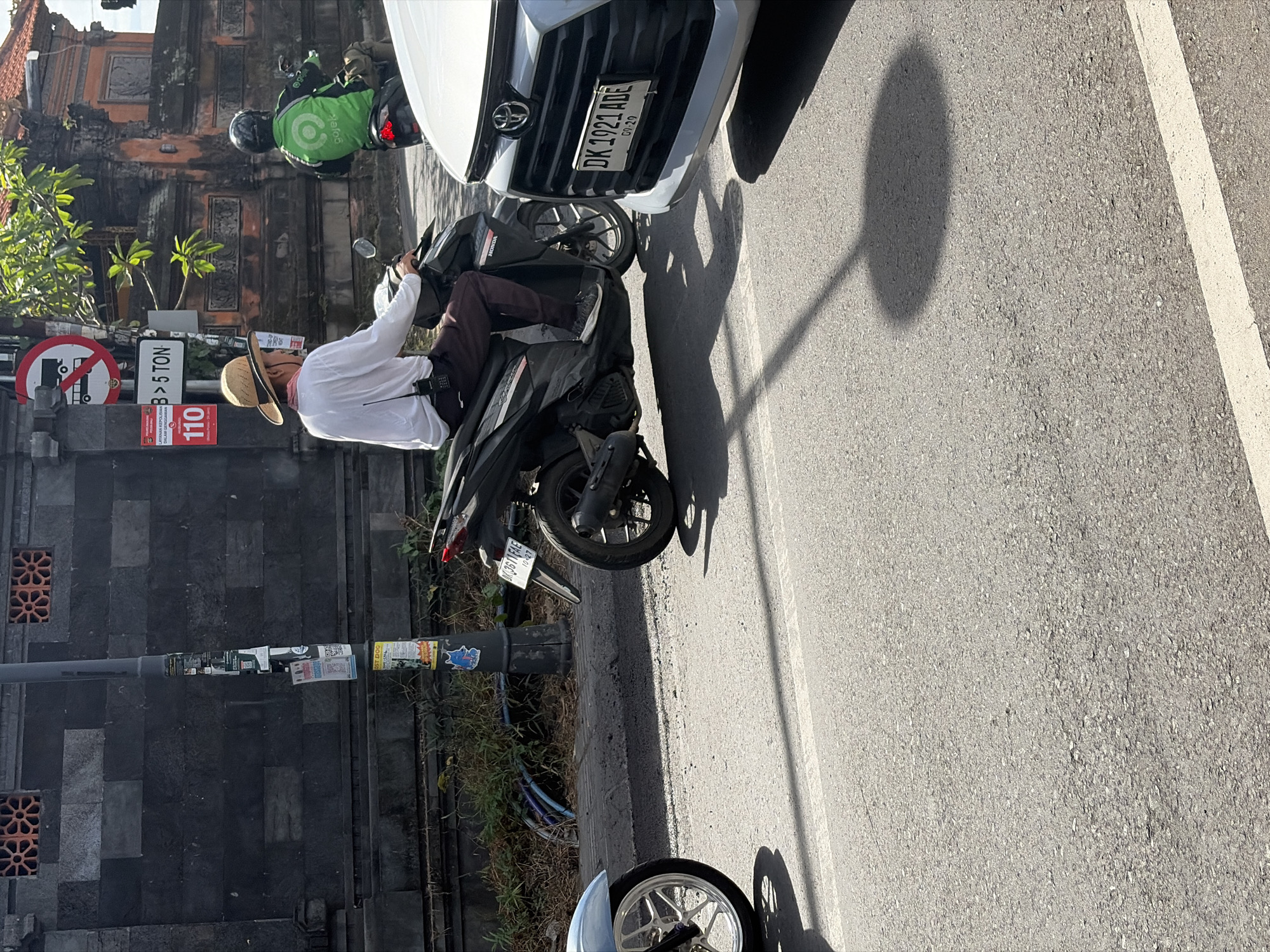 A man wearing a straw hat and white long-sleeved shirt, dark pants, and gray sneakers is riding a black Honda scooter. He has a small black radio attached to his belt. Behind him, another person on a scooter wearing a green Gojek jacket is visible. The scooters are on a paved road. To the left, the front wheel of a silver car is visible. In the background, a wall with decorative openings and a street lamp covered in stickers are present. Traffic signs are also visible on the street lamp, including one prohibiting trucks over 5 tons. The scene appears to be in North Kuta, Indonesia.