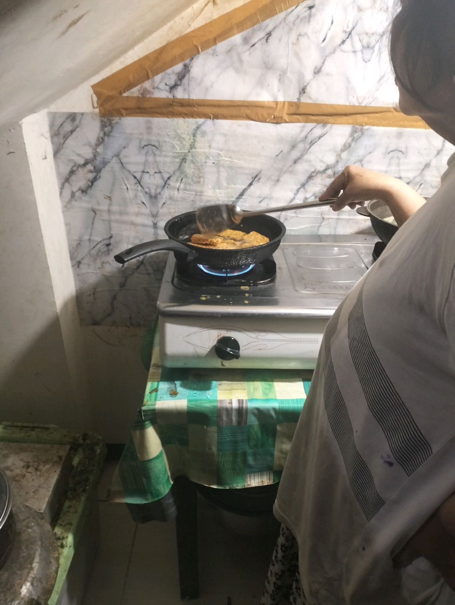 A person is standing in front of a stove in a kitchen, cooking food in a frying pan. The person is wearing a light-colored shirt. The kitchen appears to be in a residential area of Klecorejo, Indonesia, with a marble-patterned backsplash and a checkered tablecloth. The overall scene suggests a domestic cooking activity.