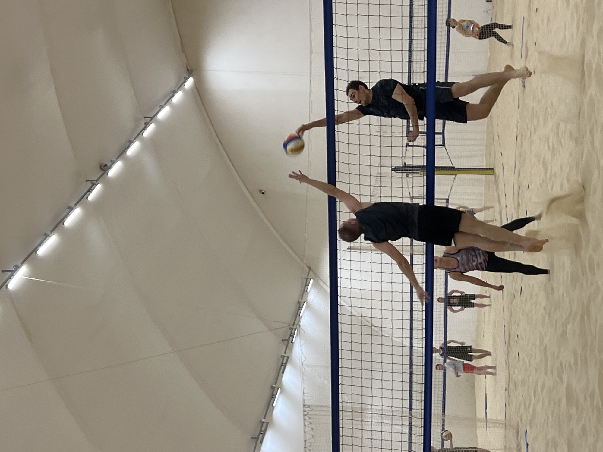 The image shows an indoor beach volleyball game. Two men are in mid-air, reaching for the volleyball over the net. Several other people stand on the sand in the background. The game appears to be taking place inside a large tent structure with lights along the ceiling. The environment is a sand court indoors. The exact location within Saint Petersburg, Russia cannot be determined solely from the image.