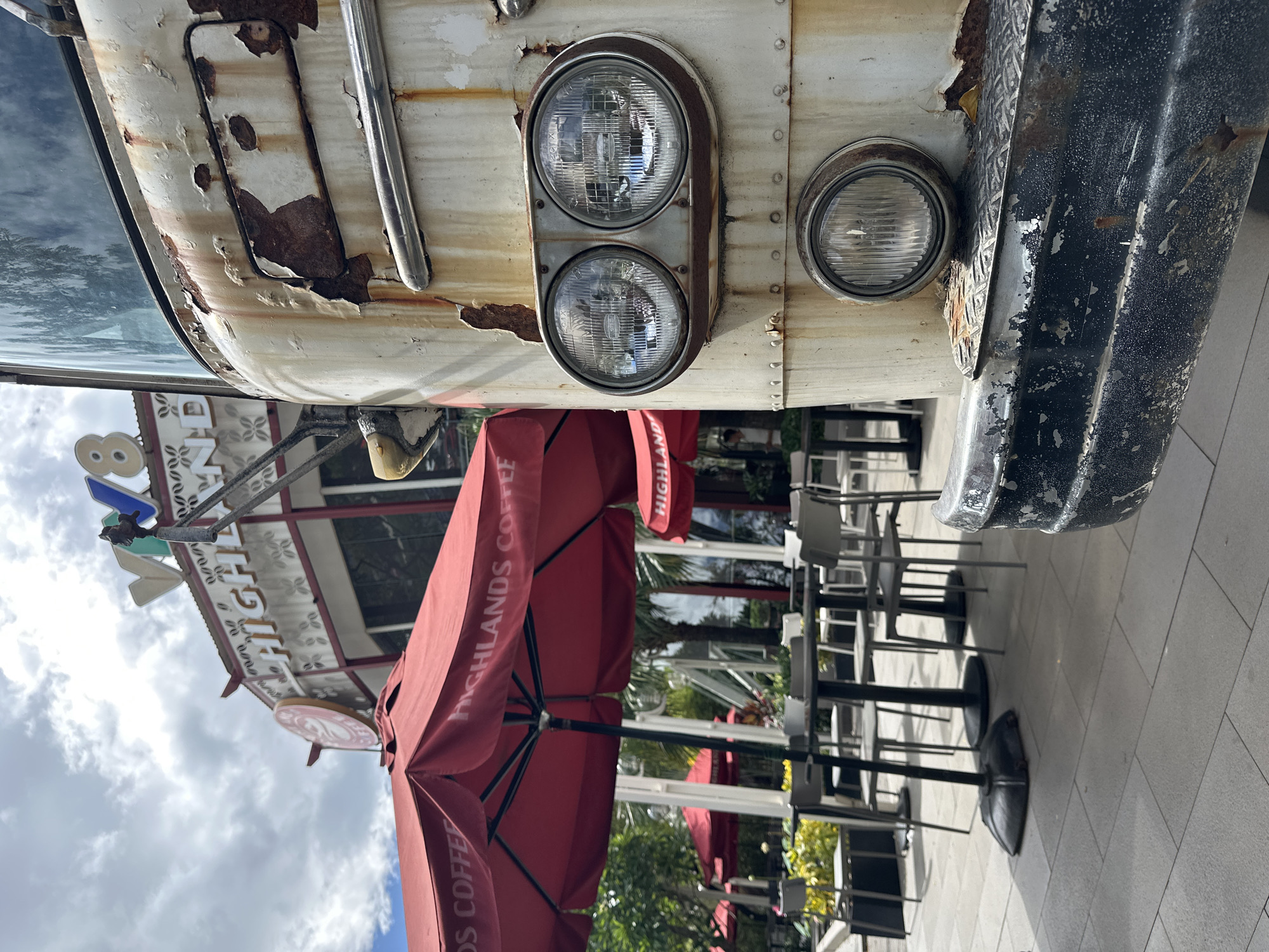 The image depicts an outdoor cafe setting in Đà Nẵng, Vietnam, centered around an old, weathered white vehicle. The front of the vehicle, possibly a bus or food truck, dominates the left side of the frame, showing extensive paint peeling and rust. It features a distinctive headlight arrangement: two round headlights are vertically stacked on the left side, and a single round headlight is visible on the right. A dark, textured bumper is partially visible on the far right of the vehicle.
In the midground, several large, red outdoor umbrellas are open, providing shade for an array of black-based tables and chairs arranged on a paved patio. The words "HIGHLANDS COFFEE" are prominently displayed in white lettering on the fabric of the umbrellas, identifying the establishment as a branch of the Vietnamese coffee chain. Further back, a building structure is visible, featuring a sign that also reads "HIGHLANDS." Green foliage from trees and bushes can be seen beyond the seating area.
The scene is set during the daytime under a partially cloudy sky. The rusted condition of the vehicle suggests a vintage or deliberately distressed aesthetic for the cafe. No people are visible, indicating a quiet moment at the outdoor seating area.