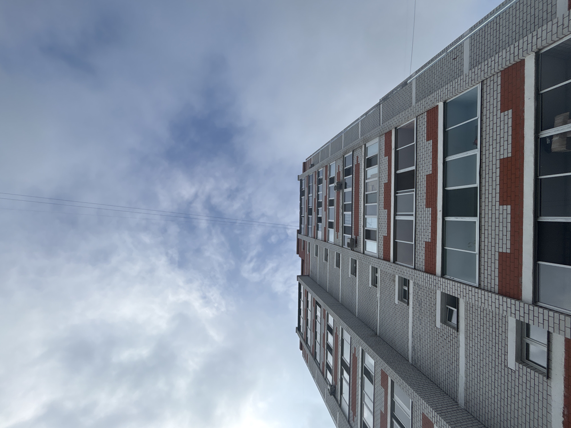 The image shows a tall, multi-story apartment building viewed from below, with the sky filling the upper portion of the frame. The building has a facade of light grey bricks and red brick accents. Numerous windows, many of which appear to be closed, are visible on each level. Electrical wires stretch across the sky from left to right. The sky is cloudy and overcast. The scene is characteristic of a residential area in Blagoveshchensk, Russia.