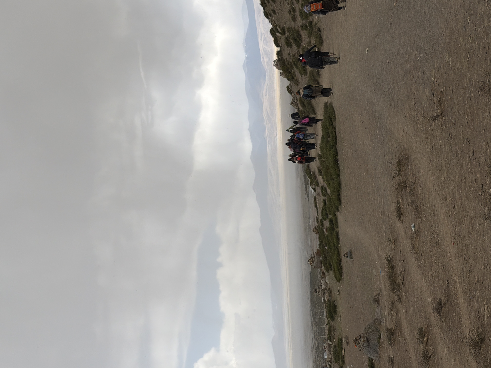 The image captures a wide, arid landscape under a heavily overcast sky. A group of approximately two dozen people are visible, some walking and others riding pack animals, likely donkeys or mules, along a winding dirt path. The path traverses a dusty, rocky hillside characterized by sparse, low-lying green vegetation and scattered dry bushes.
In the background, multiple layers of mountains recede into the distance, appearing hazy and diffused, possibly due to atmospheric dust or mist. The sky is entirely covered by thick, grey clouds, creating a soft, diffused light across the scene, indicating daytime without direct sunlight.
The individuals are mostly dressed in dark clothing, and several appear to be carrying backpacks, suggesting a trek or journey through this rugged terrain. At least two people are clearly seen riding animals, utilizing them for transport. The overall environment appears challenging and remote. No visible text is present in the image. Given the context, this scene depicts travel in a rural, mountainous area, potentially near Parga town, China.