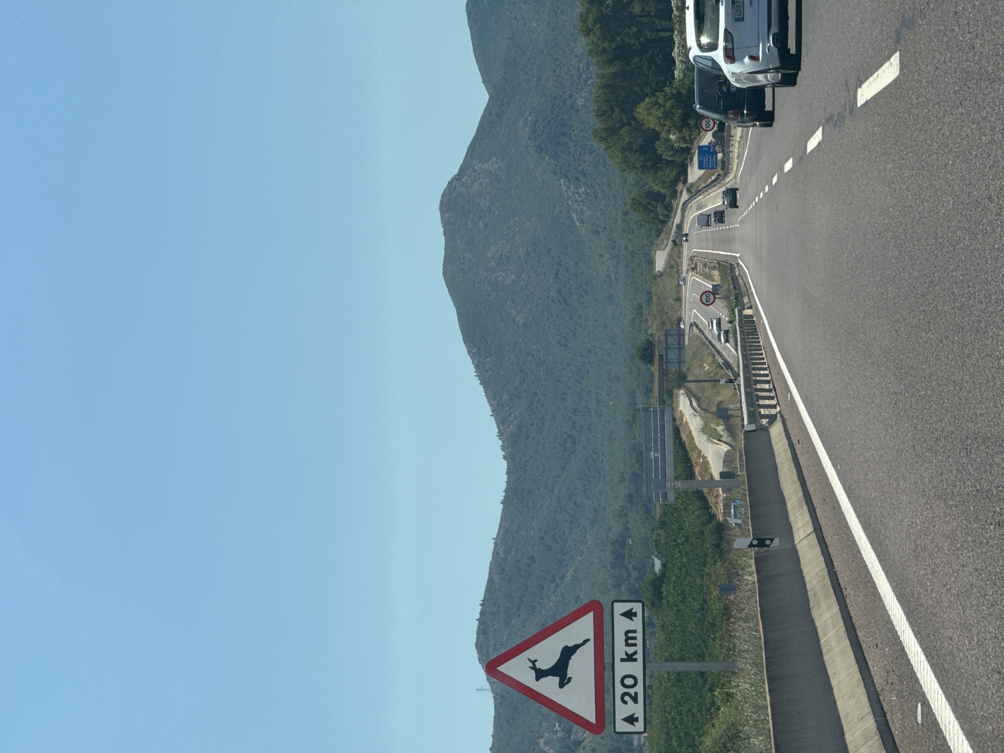 The image depicts a view from a vehicle traveling on a highway in Xàtiva, Spain. The road is paved with asphalt and marked with white lines. To the left of the road, there is a concrete barrier and a triangular warning sign indicating deer crossing, with "20 km" written below it. Beyond the barrier, there are green fields and hills covered with vegetation. In the distance, a range of mountains forms the backdrop under a clear blue sky. Several vehicles are visible on the highway, traveling in the same direction as the viewer. The scene appears to be captured during daylight.