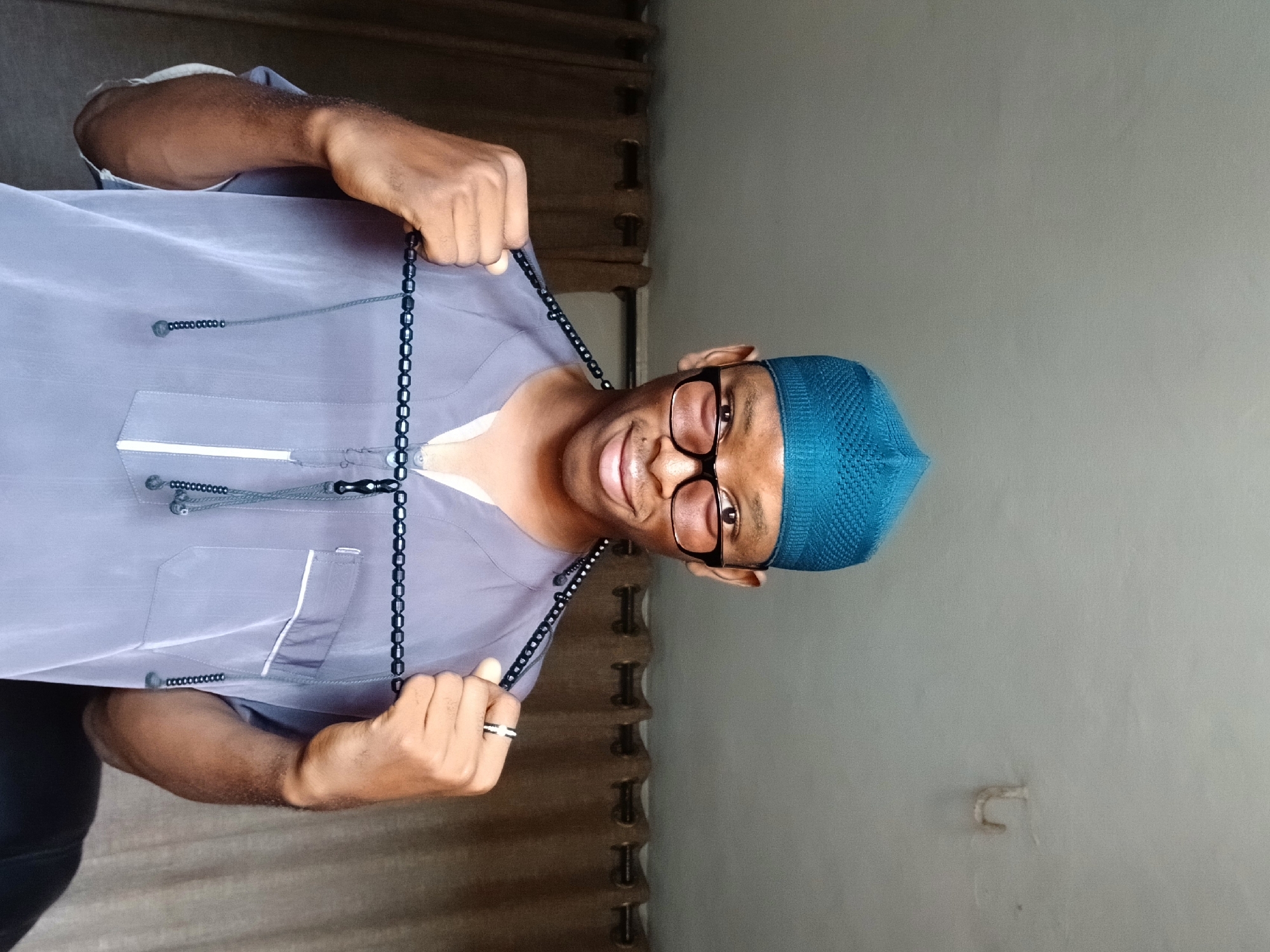 A man wearing a blue kufi and glasses is smiling and holding a black tasbih (prayer beads) in front of him. He is wearing a light gray tunic-style shirt. The background is a neutral-colored wall, and there are curtains visible behind him. The location is likely indoors and could be in Lagos, Nigeria, given the subject's attire and the likelihood of the photo being taken where similar cultural and religious expressions are found.