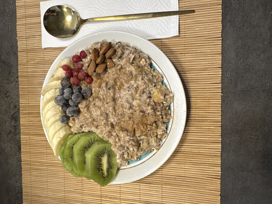 A plate of oatmeal decorated with banana slices, blueberries, red currants, almonds, and kiwi slices rests on a woven mat. A gold-colored spoon lies across a white napkin above the plate. The oatmeal itself appears to be sprinkled with cinnamon. The overall arrangement suggests a healthy and aesthetically pleasing breakfast or meal.