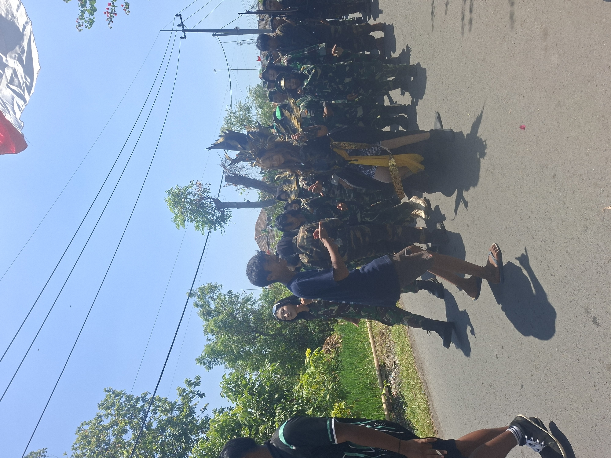 In Tongaswetan, Indonesia, a group of people dressed in camouflage uniforms and a person in a decorative headdress are participating in a procession along a paved road. The individuals in camouflage are marching in formation, some holding what appear to be rifles. The person in the elaborate headdress, adorned with feathers and gold accents, is positioned at the front of the group, walking alongside a person in a dark blue t-shirt. The environment is characterized by a clear blue sky, lush green trees and vegetation lining the road, and power lines overhead. A flag, partially visible at the top of the image, suggests a festive or commemorative event. The road is casting shadows from the procession and surrounding trees.