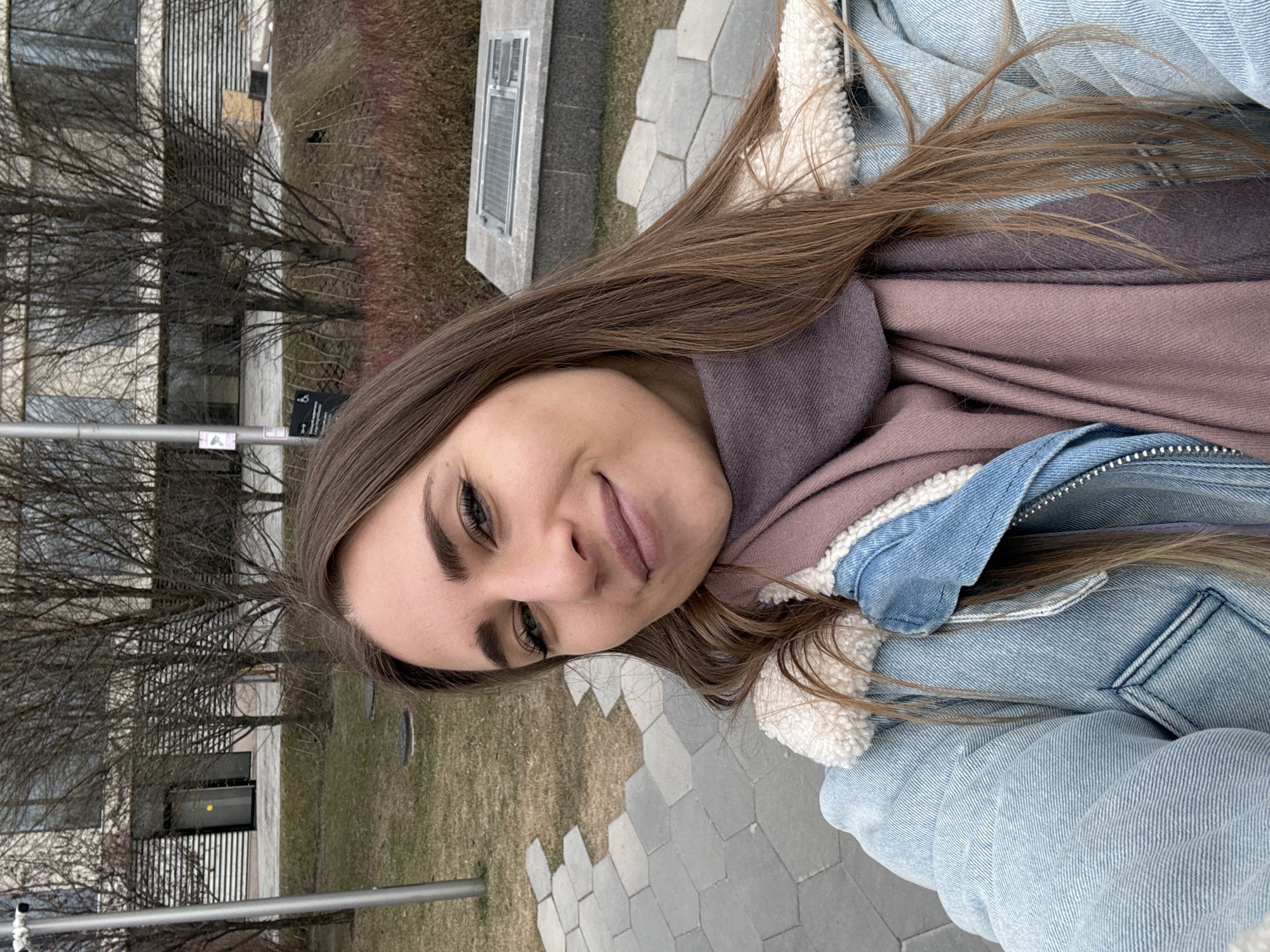 A female individual is depicted in a close-up outdoor selfie, smiling directly at the camera. She possesses long, light brown hair and is dressed in a light blue denim jacket with a cream-colored sherpa lining visible at the collar and shoulders. A two-toned scarf, appearing in shades of mauve and dark purple/gray, is wrapped around her neck.
The background details an urban park landscape. Hexagonal grey paving stones are present in the foreground, transitioning into patches of dry, sparse grass and areas of reddish-brown vegetation. Numerous deciduous trees with bare branches are scattered throughout the mid-ground, suggesting a late winter or early spring season. To the left, a slender metallic pole displays a white sign featuring a surveillance camera icon and a wheelchair accessibility symbol with Cyrillic text. Behind the trees, a modern building with a light-colored, horizontally-slatted facade and large windows is visible, including a brightly illuminated entrance. To the right, a rectangular concrete structure topped with a metal grate rests on a low earthen mound.
The location is identified as Zaryadye Park in Moscow, Russia, based on its distinctive paving, contemporary architecture, and landscaped features.