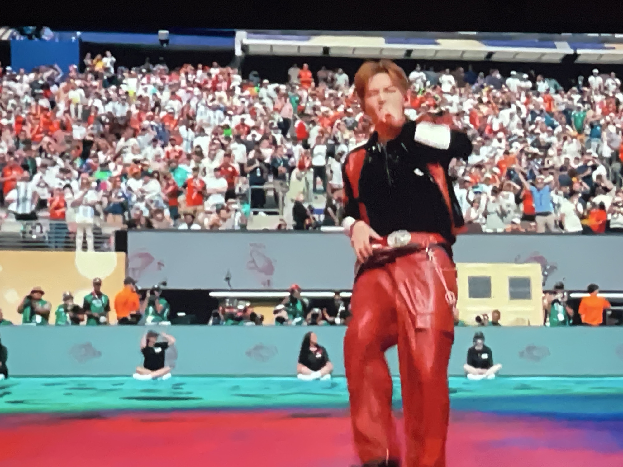 A wide-angle, medium-shot photograph shows a performance on a brightly lit stage in front of a large stadium crowd. The performer in the foreground is a young male with blond hair, wearing a black shirt with red accents and red leather pants. He is in motion, with his mouth open as if singing or shouting, and his right arm is raised. The crowd in the background is dense and appears to be watching the performance. Several individuals are visible in the middle ground, some appearing to be dancers or performers on the stage, one of whom is sitting cross-legged on a bright teal platform. The stage floor is a gradient of bright colors, transitioning from teal to pink. The overall atmosphere suggests a large-scale entertainment event, possibly a concert or festival.