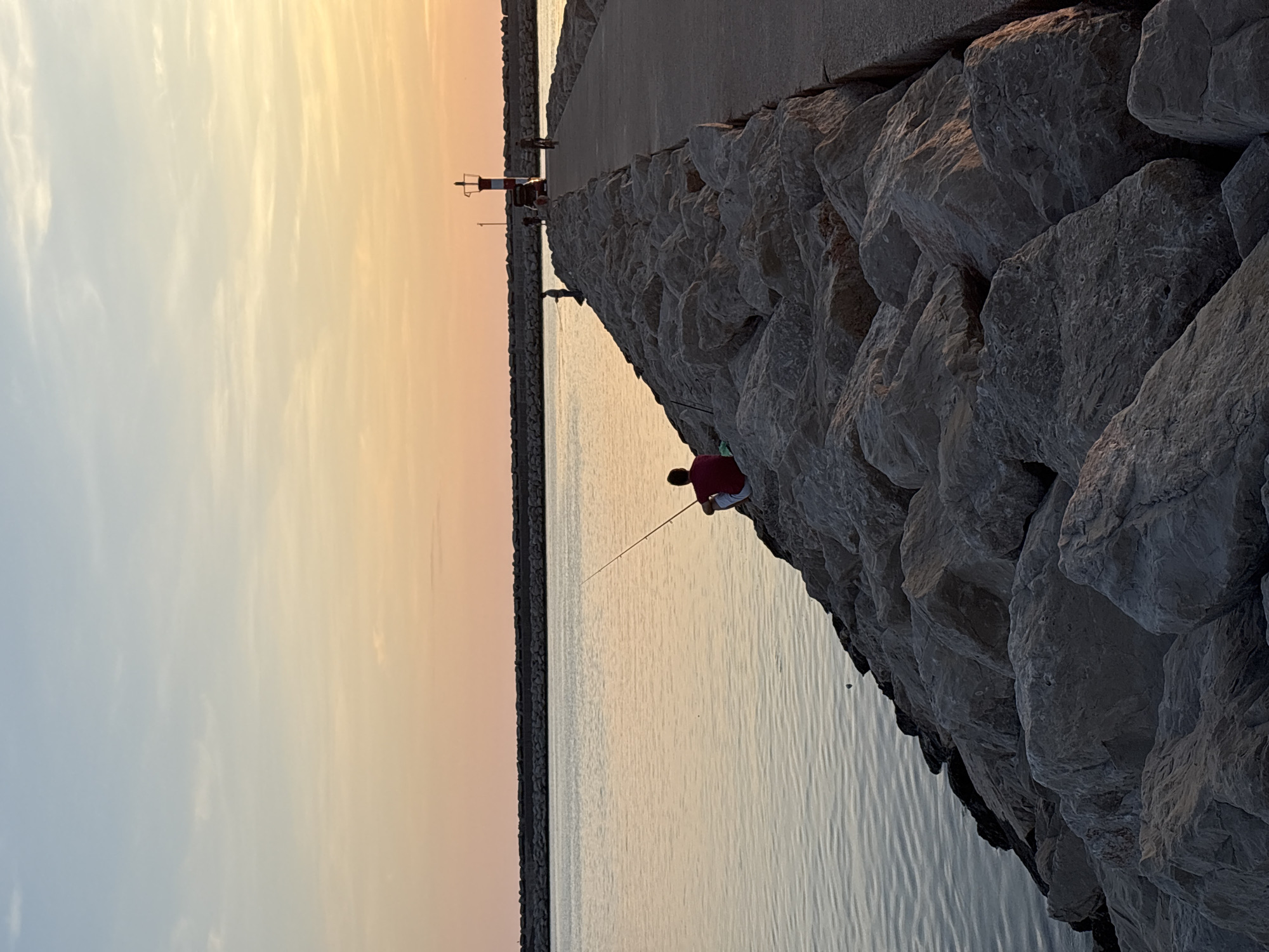 A wide-angle, outdoor shot depicts a fishing scene at sunset on a rocky pier, likely in Bar, Montenegro. The sky is a gradient of orange and pale yellow, transitioning to a light blue towards the top. A breakwater of large, grey rocks slopes down into calm water that reflects the sky's colors. A person is sitting on the rocks, wearing a red shirt and white pants, fishing with a rod that extends out over the water. Further down the pier, there is a small structure, potentially a lighthouse or navigation aid, marked with red and white. The pier is built of concrete on top of large rocks. The image suggests a peaceful evening scene.