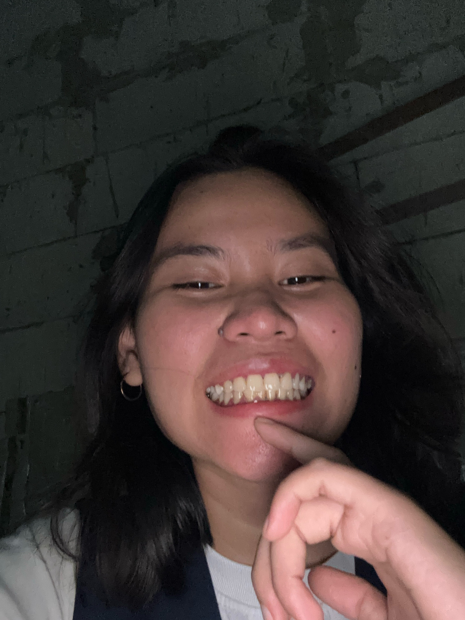 A young woman of Asian descent is taking a selfie in a dimly lit indoor environment. She has dark, shoulder-length hair and is wearing a white t-shirt under a dark blue garment. She is smiling, revealing her teeth, and has her right index finger touching her chin. A small hoop earring is visible in her left ear, and a nose stud is present on her right nostril. The background consists of a textured wall that appears to be made of concrete blocks, with some horizontal wooden beams visible at the top. The lighting is dim, casting shadows on the wall and highlighting the woman's face. There is no indication of her specific location within East Jakarta, Indonesia, beyond it being an indoor space.
