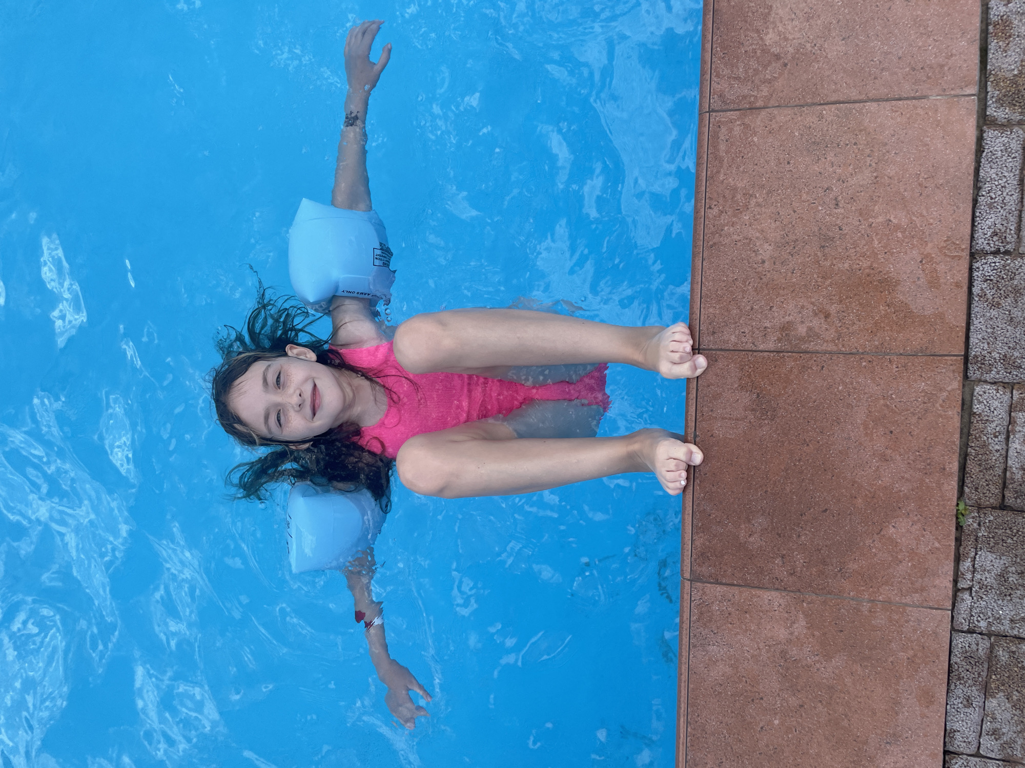 A young girl with dark, curly hair is floating on her back in a bright blue swimming pool. She is wearing a bright pink swimsuit and light blue arm floaties. Her arms are outstretched, and her legs are bent at the knees. She is smiling at the camera, with a happy and relaxed expression. Her hair is wet and spread out in the water.
The pool has a tiled edge, with reddish-brown tiles on one side and a darker, rougher surface with small stones on the other. The water is clear and reflects the sky, creating shimmering patterns on the surface. The perspective is from above, looking down at the girl in the water.
There is some text visible on the arm floaties, but it is too blurry to read clearly. The scene appears to be taking place on a sunny day, as indicated by the bright light and reflections on the water. The overall impression is one of joy and carefree enjoyment of a summer day.