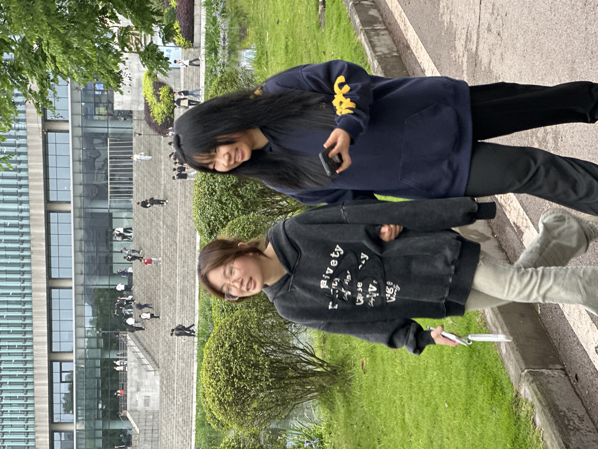 Two young women stand on a paved walkway beside a grassy area with bushes, in front of a multi-story building with glass windows. Numerous people are visible on the steps leading up to the building's entrance. The woman on the left wears a grey hooded sweatshirt, light-colored jeans, and glasses, and she holds a white object in her left hand. The woman on the right wears a dark blue sweatshirt with yellow lettering on the sleeve and black pants, and she holds a black smartphone. The location is Jiangjin, China.