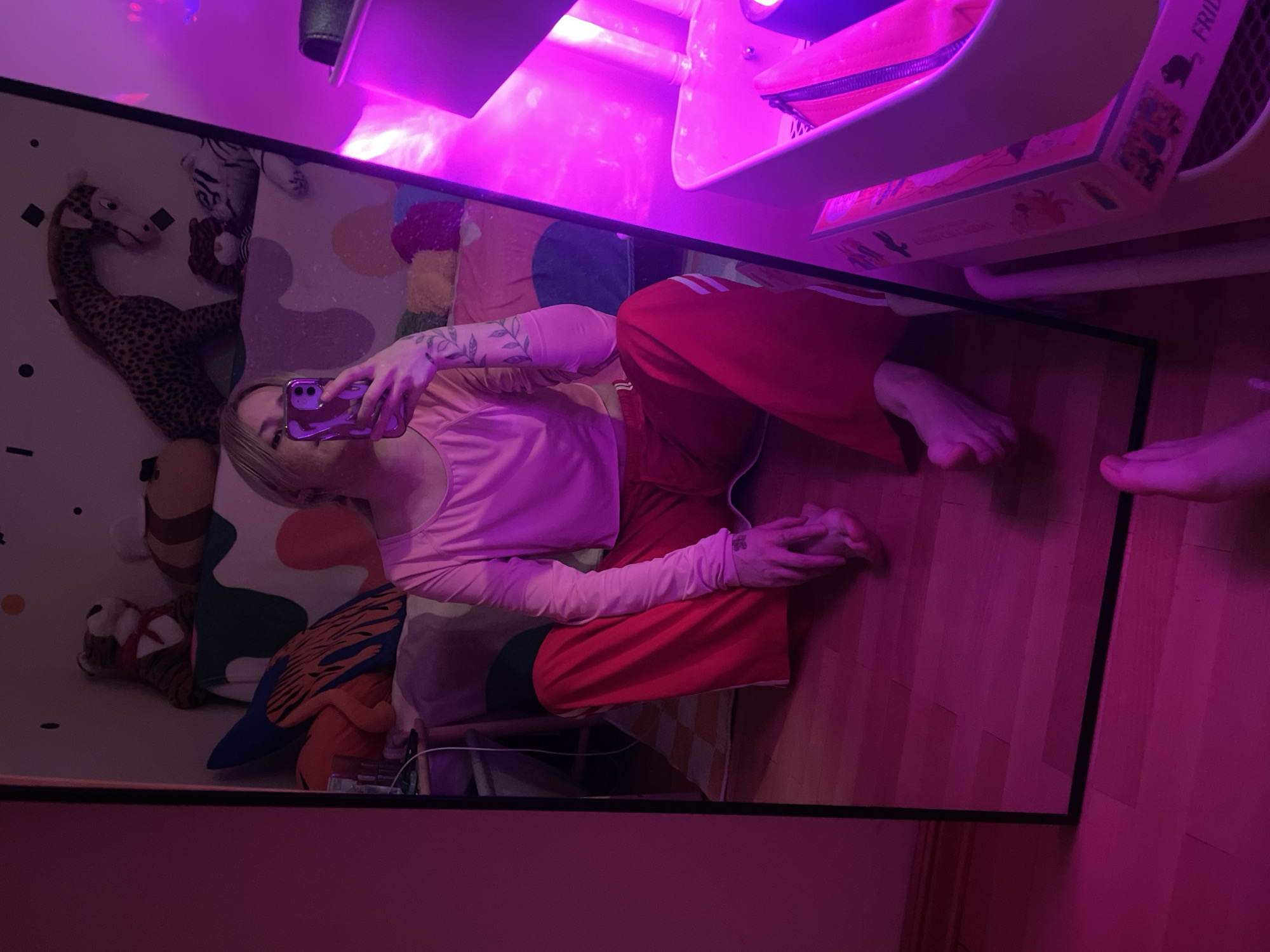 The image displays a mirror selfie taken indoors, set against a backdrop illuminated by a dominant magenta/pink light source. The scene is located in Belgrade, Serbia.
A person is lying on their side, reflected in a large mirror. They have blonde hair and are dressed in a light pink long-sleeved top and red sweatpants with white stripes running down the sides. Tattoos, possibly depicting branches or leaves, are visible on both forearms. The person holds a purple smartphone up to their face, actively taking the selfie.
Reflected in the mirror alongside the person is a section of a room. This area features a patterned rug or playmat, upon which several stuffed animals are placed, including a large, spotted, long-necked animal resembling a giraffe, and a striped animal akin to a tiger.
Outside the mirror's frame, on the right, the person's bare feet are visible on a wooden floor. In the upper right corner, a piece of furniture, possibly a desk or shelf, is partially visible, under which a box with the word "FRIDA" printed on its side can be seen. The pervasive magenta light creates a distinct, vibrant atmosphere throughout the image. The activity depicted is a casual mirror selfie, with the person in a relaxed, reclined pose.