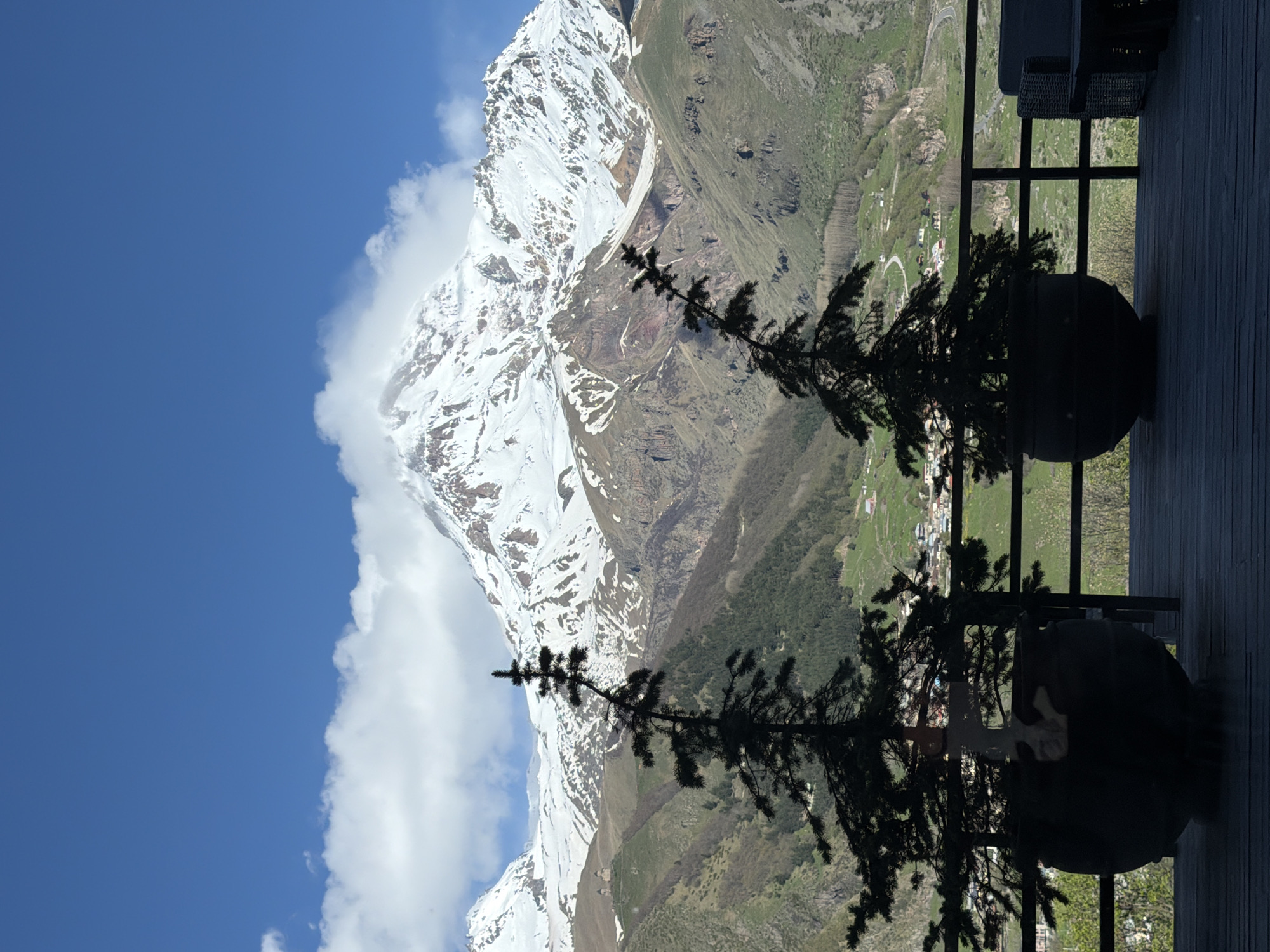 The image shows a view from a balcony in Stepantsminda, Georgia, towards a snow-capped mountain. The sky is blue with some clouds near the mountain peak. A black metal railing runs along the balcony edge. Two large, round planters are visible on the balcony, with silhouettes of small evergreen trees inside. The mountain slopes down to green hills with a small town nestled within. A winding road can be seen in the background.
