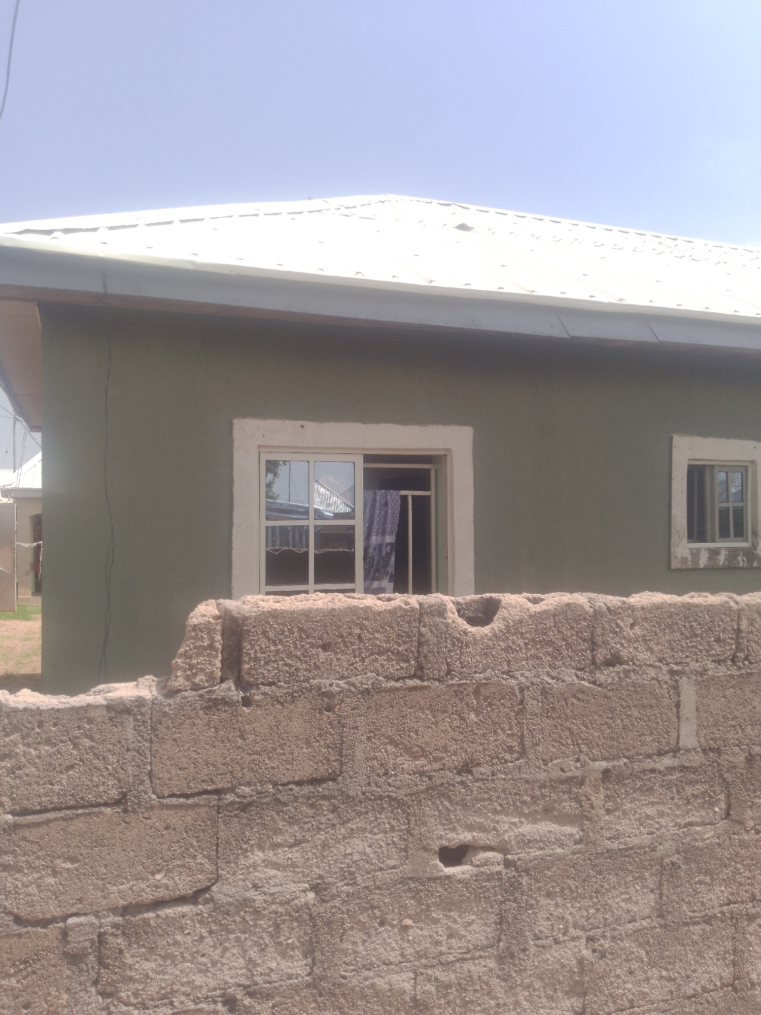The image depicts a house with a light-colored metal roof and olive green walls. In the foreground, a rough, light brown brick wall is visible, partially obscuring the view of the house. The house has at least two windows; one larger window with multiple panes is centrally located, and a smaller, similar window is situated to the right. The larger window shows a glimpse of patterned curtains inside. The sky above is a clear, light blue, suggesting a bright, sunny day. The overall scene appears to be an outdoor setting, possibly residential. There are no people or discernible activities taking place. The location cues like the building style and the brick wall suggest a setting in Jauro Yinu, Nigeria. There is no visible text in the image.