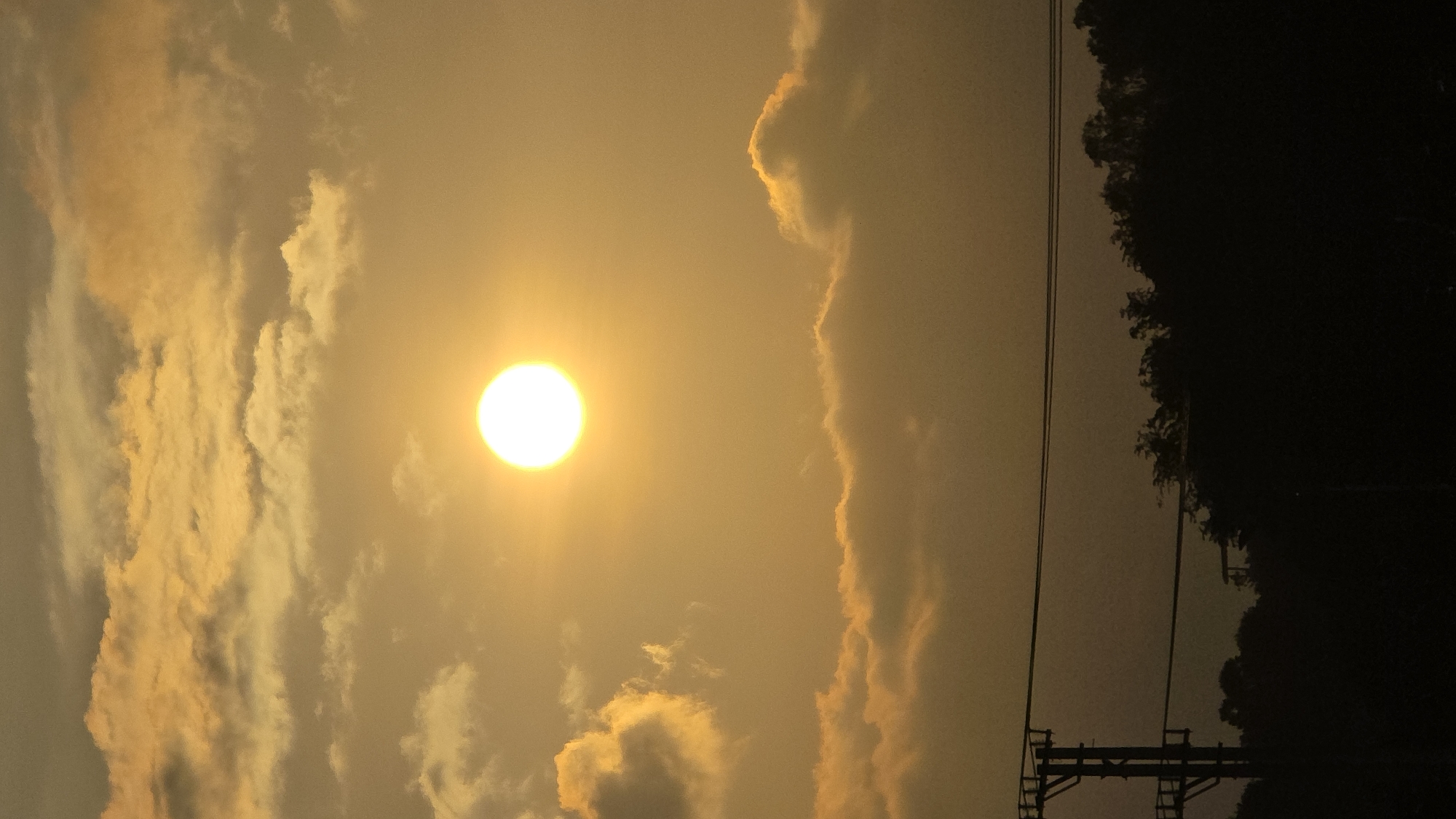 The image captures a bright sun in a hazy, yellowish sky, with various clouds scattered across the scene. To the right of the sun are some vertically oriented, elongated clouds with bright edges. Farther to the right, silhouetted trees and an electrical power pole stand against the sky. The photo seems to be taken during sunset or sunrise due to the golden hue. Nha Trang, Vietnam is not discernible from the context of the image, there is no way to ascertain the city from this photo.