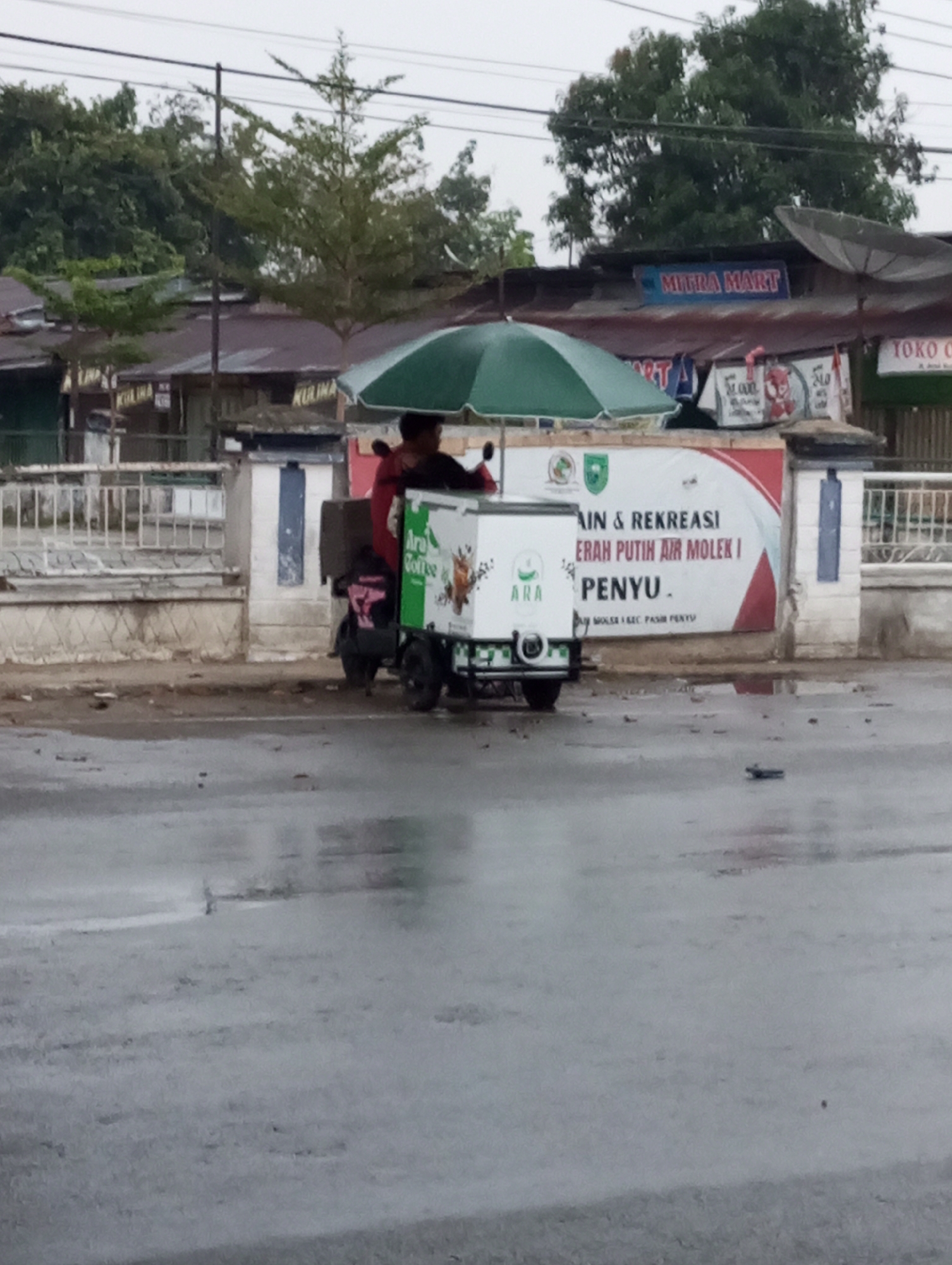 The image depicts a street scene in Air Molek, Indonesia, on a wet day, indicated by the reflective surface of the asphalt road. A vendor is operating a mobile cart, likely for selling drinks or ice cream, which is attached to a scooter. The cart is white and green, featuring branding that includes the word "Ara" and an image of what appears to be a coffee cup. The vendor, wearing a red garment, is seated on the scooter. Behind the cart, a large banner with red and white graphics and text is displayed. The text on the banner includes "TAIN & REKREASI," "ERAH PUTIH AIR MOLEK I," and "PENYU." This suggests the banner is advertising an event or location related to recreation in Air Molek. Further in the background, various buildings with corrugated metal roofs are visible, along with trees and a satellite dish, hinting at a commercial or semi-urban environment. Signs for "MITRA MART" and "TOKO C" are also discernible on some of the buildings, indicating the presence of local businesses. The overall atmosphere is subdued, possibly due to overcast weather.