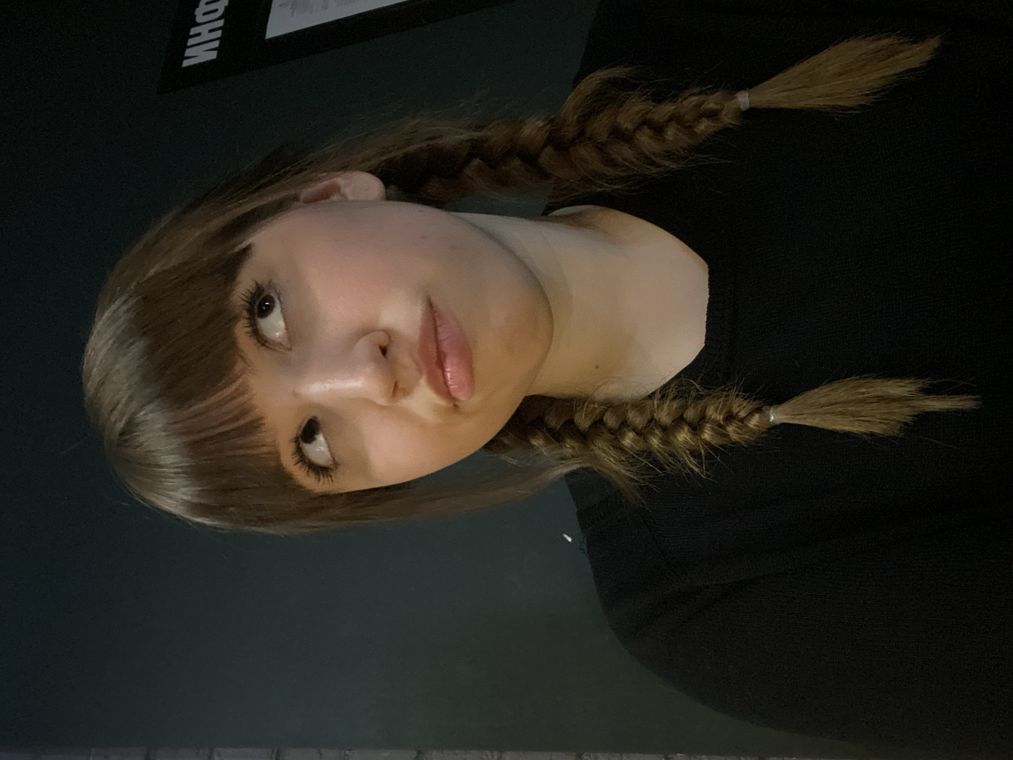 The image is a portrait of a young woman, rotated 90 degrees counter-clockwise. She has light brown hair, featuring bangs across her forehead and two distinct braids on either side, each secured with a light-colored elastic band. Her head is tilted, and she looks upwards and slightly to her right with a thoughtful or contemplative expression. Her fair skin is illuminated, highlighting her full, light-tinted lips and prominent eyelashes. She is wearing a dark, likely black, top or sweater. The background is dark and largely out of focus, indicating an indoor setting with low lighting. In the upper left portion of the frame, partially visible and upside down, are Cyrillic characters that appear to spell "ИНФО" (INFO). The scene is set in Chaikovsky, Russia.