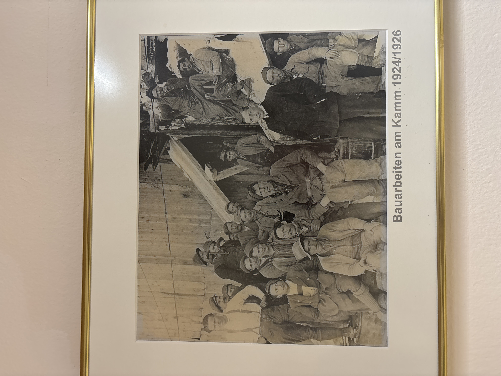 This is a black and white photograph of a group of approximately 20 men posing outdoors, likely at a construction site given the background which features rough wooden structures and debris. The men are dressed in what appears to be work attire, with many wearing hats and suspenders. Some men are seated, while others stand, with the majority looking towards the camera. In the foreground, one man is dressed in a suit, appearing to be in a supervisory role. The photograph is framed and mounted on a wall, with a printed caption below it that reads "Bauarbeiten am Kamm 1924/1926". The text below the image indicates that the location is Ehrwald, Austria.