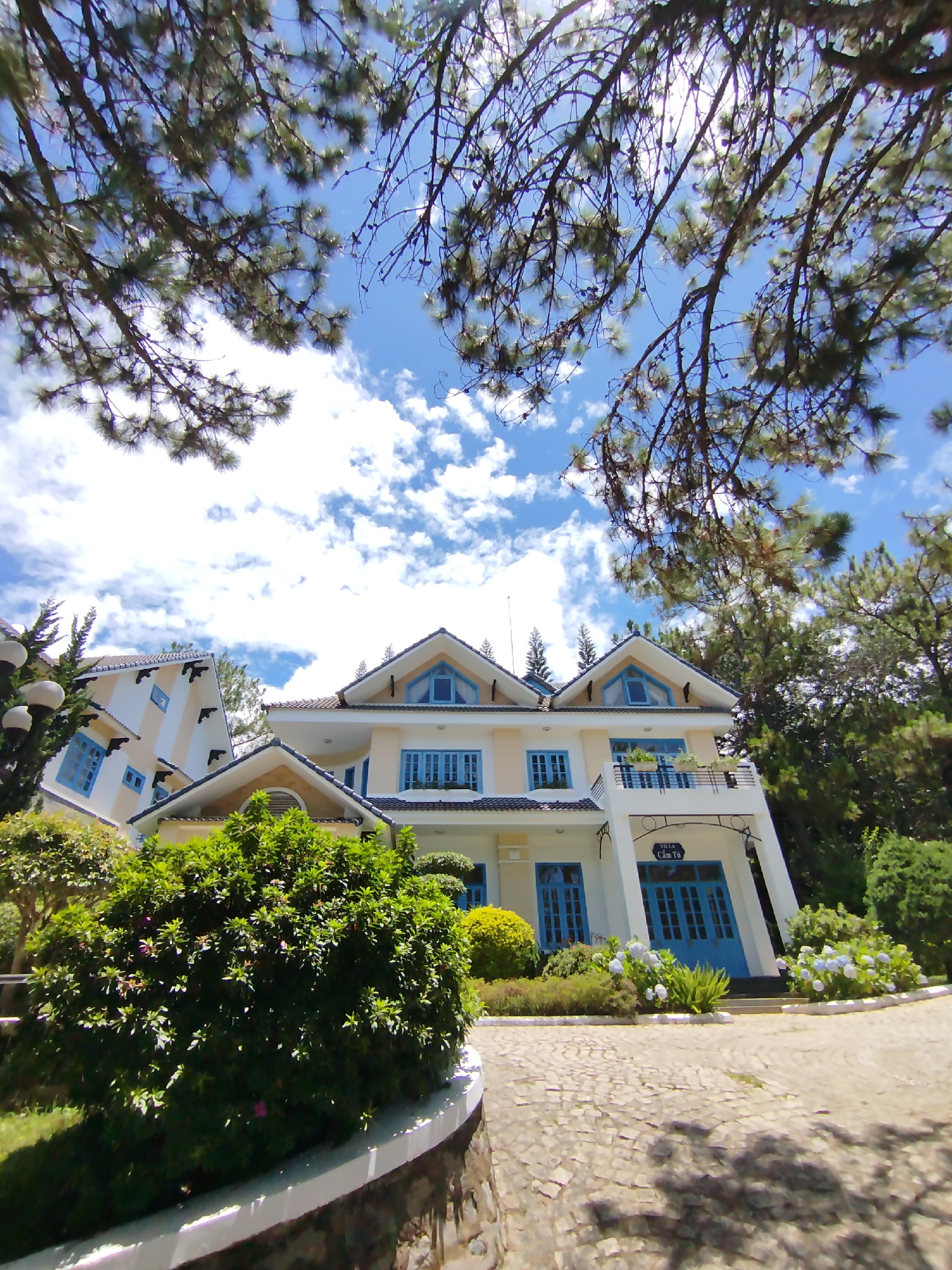The image shows a two-story, pale yellow building with striking blue window frames and doors, nestled among lush greenery and under a sky dotted with white clouds. The building has multiple gabled roofs with small, triangular windows at their peaks. A balcony is visible on the second story. A second, similarly styled building is partially visible to the left. A large, rounded bush dominates the foreground. The ground is paved with a stone-like material. Branches of pine trees frame the top of the image. The scene is identified as being in the city of Lâm Viên - Đà Lạt, Vietnam, based on the sign on the building reading "Villa Cam Tú". No people are visible.
