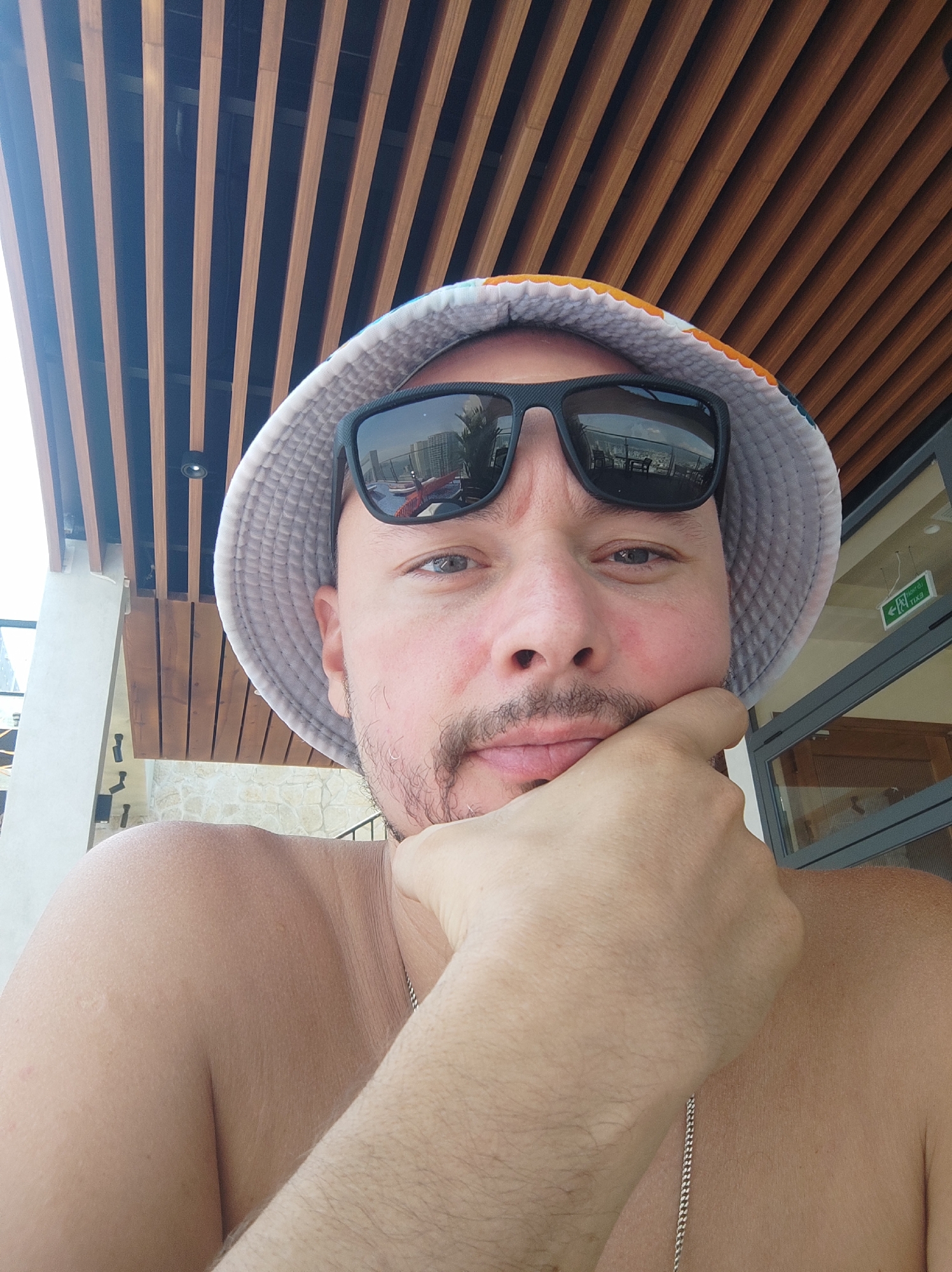 A man wearing a bucket hat and sunglasses rests his chin on his hand, looking at the camera. He is shirtless, revealing a silver chain necklace. The background features a wooden slatted ceiling and a glass door with an exit sign. The man's reflection in his sunglasses shows a swimming pool and buildings. The scene appears to be outdoors in a resort or hotel setting in Nha Trang, Vietnam, possibly near a pool.