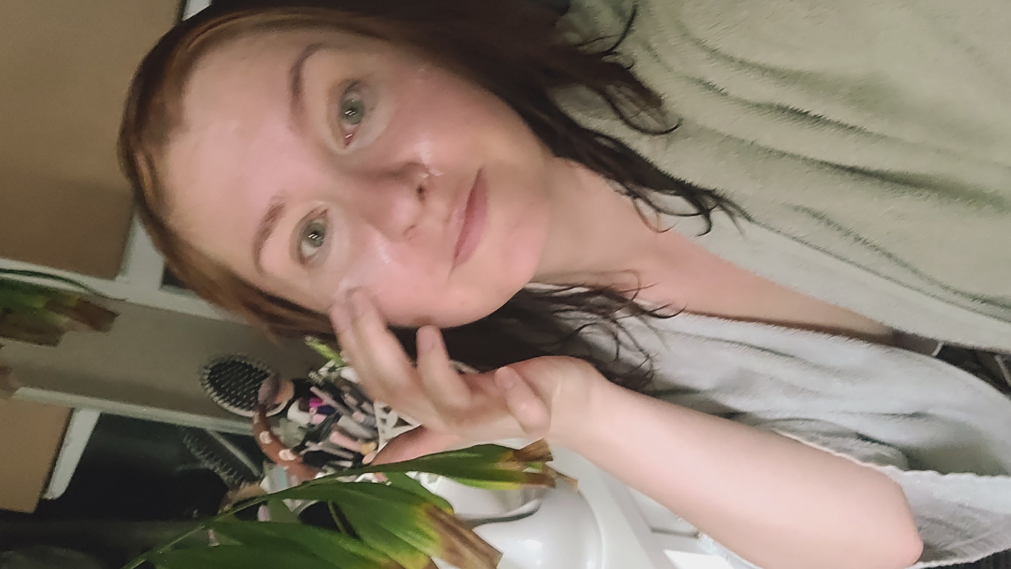 A pale-skinned woman with auburn hair is applying cream to her face. She is wearing a light gray bathrobe. Her eyes are green and she has fair skin. In the background, a green plant is visible, along with some makeup brushes and other items on a shelf. The lighting is dim, suggesting it is either early morning or evening.