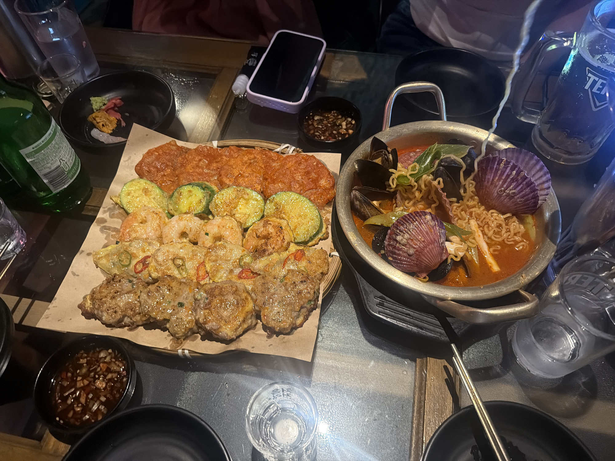 The image depicts a table laden with Korean food in a dimly lit environment, likely a restaurant in Seoul, South Korea. Notable items include a large pot of seafood ramen with mussels and scallops, a platter of assorted savory Korean pancakes (jeon) including meat, zucchini, and shrimp varieties, and small bowls of dipping sauce. A green bottle, likely soju, and glasses of alcohol suggest a social gathering. A smartphone is also present on the table. The setting is a casual dining scene.