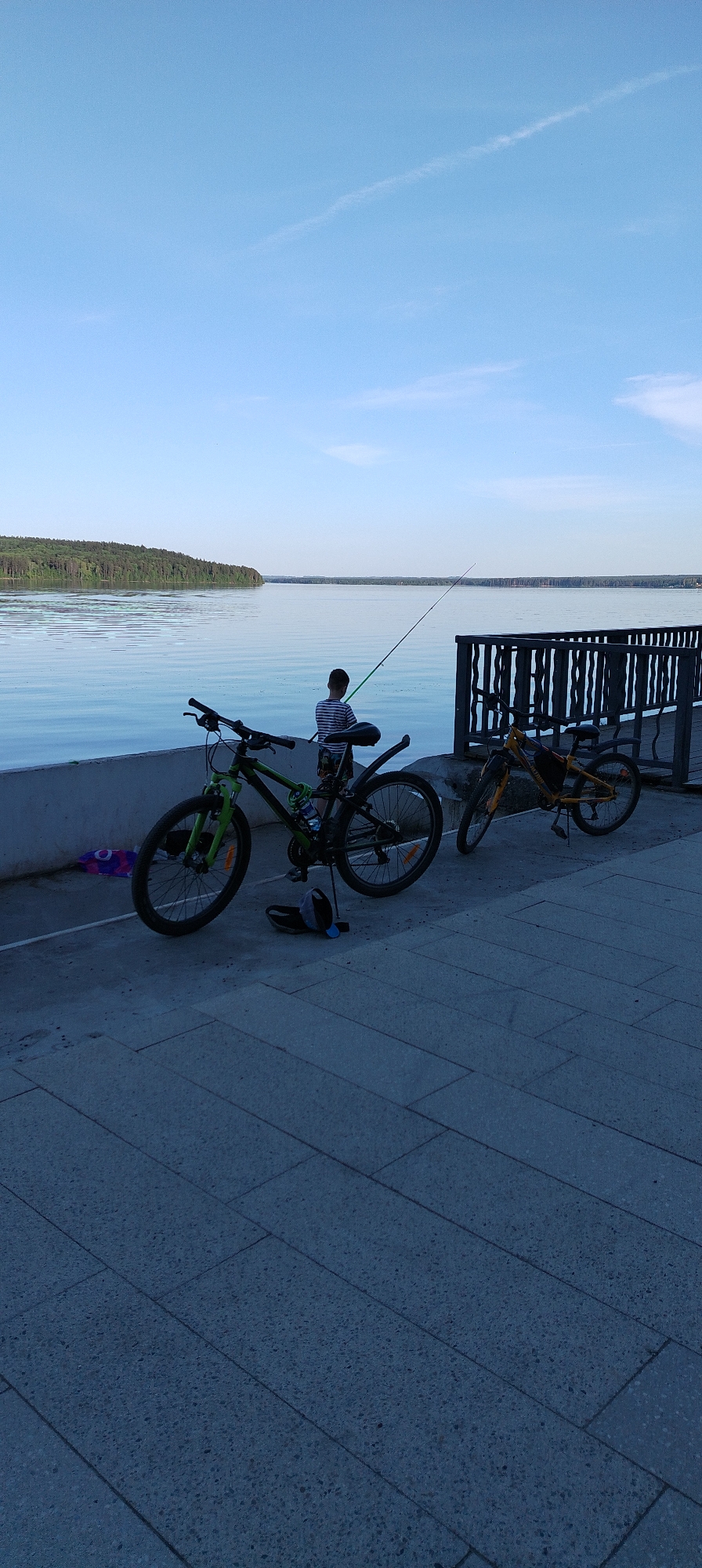 The image depicts a waterfront scene with a person fishing in the city of Chaikovsky, Russia. A body of water, possibly a lake or river, stretches into the distance with a tree-lined shore visible on the opposite bank. The sky is a clear, pale blue.
In the foreground, two bicycles are parked on a paved walkway. One bike is green and black, positioned closer to the viewer, while the other is yellow and located near a wooden railing that borders the water. A person stands near the railing, holding a fishing rod. They are facing the water.
The paved area is made of rectangular stones. The overall lighting suggests it could be dusk or dawn.