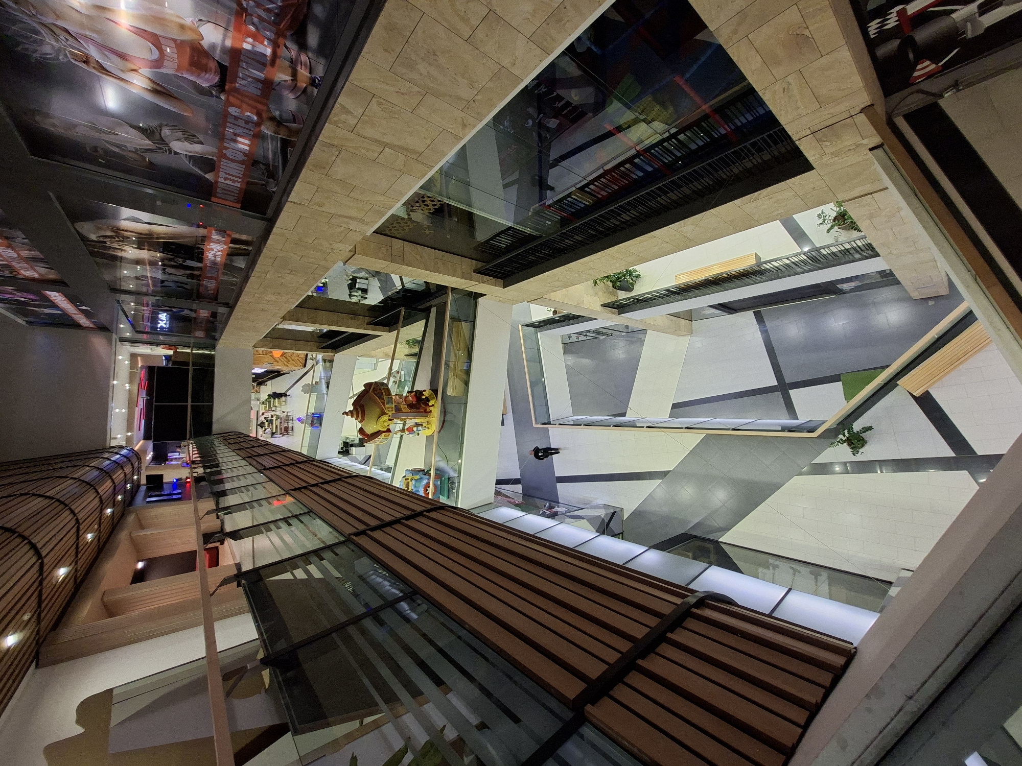 This is an interior view of a multi-level shopping center in Plovdiv, Bulgaria, taken from an elevated perspective looking down into the atrium. The atrium is characterized by a geometric floor pattern of white and gray tiles interspersed with black lines, some sections appear to be seating areas with benches. The central part of the view is the internal architecture, with different levels visible, enclosed by glass and supported by wooden structures.
The light entering the scene is a mix of natural light, most likely through a skylight, and artificial lighting that illuminates different parts of the shopping center. A carousel, possibly a children's ride, is visible through the structure. The higher levels feature decorative elements and advertisements. The image is taken from an angle that suggests the observer is positioned on one of the upper floors, looking down into the heart of the commercial space.