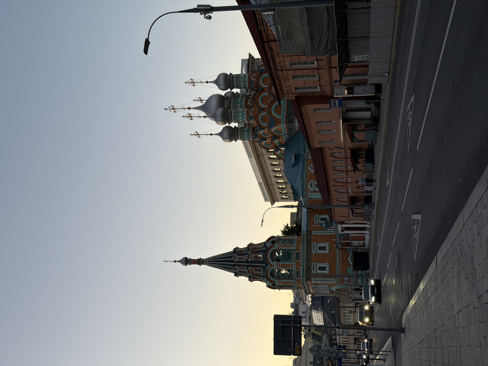 The image depicts a street scene in Moscow, Russia, featuring a prominent Orthodox church with a tall, ornate bell tower and several onion domes. The architecture suggests a historical or religious site. A paved road runs in front of the church, with cars driving on it. The sidewalks are also paved, and there are trees and streetlights visible. The sky is clear and light, indicating either early morning or late evening. The overall atmosphere is calm and urban. The church's distinctive orange and teal color scheme and intricate detailing make it the focal point of the image. There are a few pedestrians visible on the sidewalk near a bus stop.