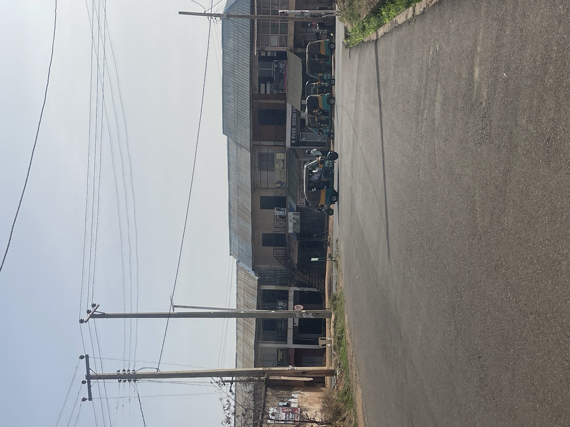 The image captures a street scene in Jos, Nigeria, looking down a paved road. On the left, a row of multi-story buildings with corrugated metal roofs and visible windows line the street, suggesting commercial or mixed-use purposes. Several green and yellow "Keke Napep" auto-rickshaws, a common form of public transportation, are parked along the storefronts. One Keke Napep has "PTA 61" visible on its side, and a person can be seen inside another. Shop signage includes "A PLUS SECRETS" prominently displayed above an awning, with partial signs for "MODABO BEAUTY" and "MODABO APPAREL" visible further along the street.
Numerous wooden utility poles stand tall on the left side of the frame, supporting a complex array of overhead power and communication lines that crisscross against the sky. The sky is bright and appears slightly overcast or hazy, indicating daytime, likely morning or afternoon.
To the right, the paved asphalt road extends into the distance, appearing relatively clear. A low curb borders the road, beyond which a strip of green grass and foliage is visible. The scene conveys a glimpse into the daily urban environment and infrastructure of a Nigerian city.