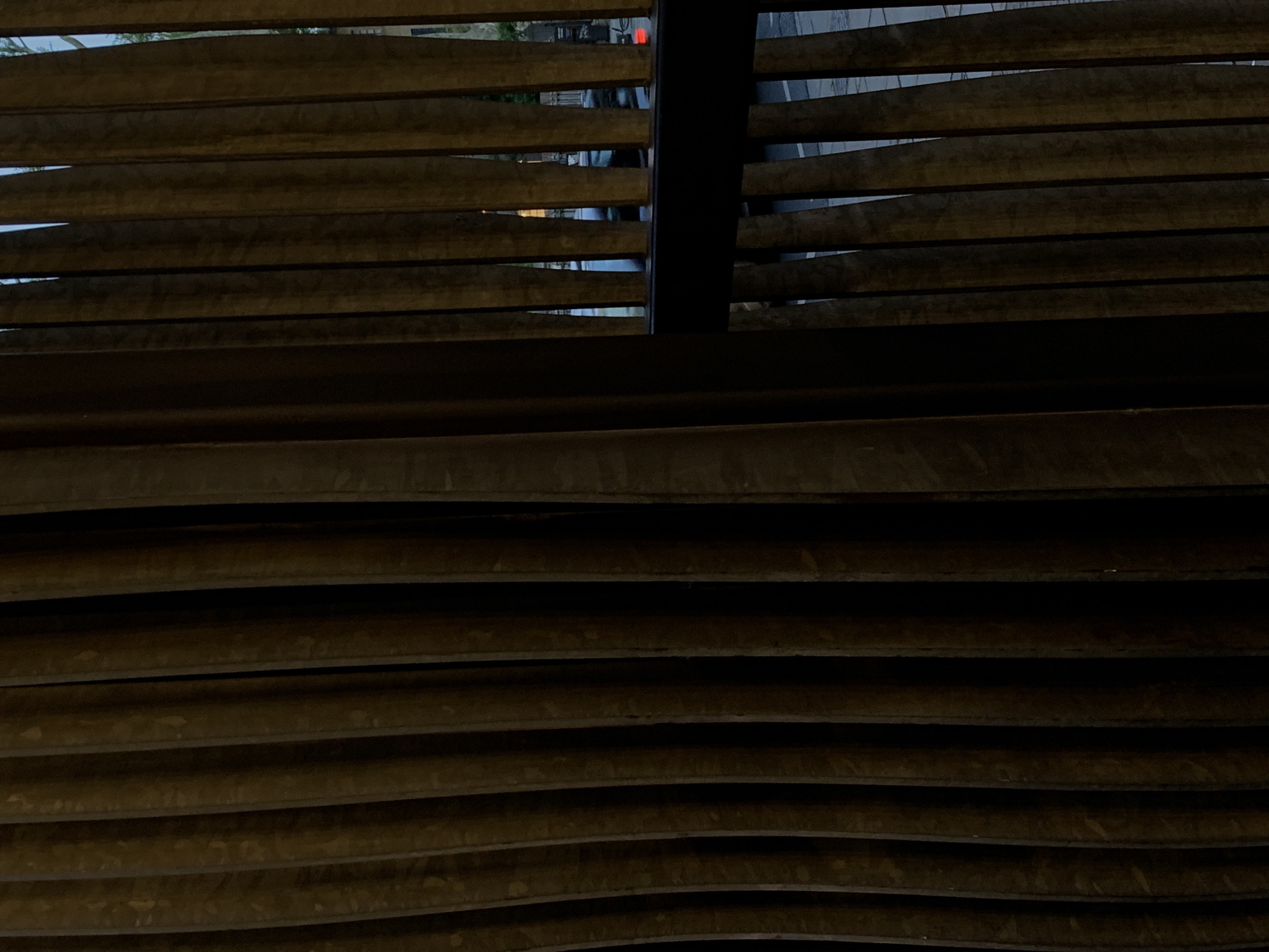 The image presents a view seen through a series of dark, horizontal slats, likely part of a louvered screen, blinds, or an architectural facade. These slats, appearing to be made of dark wood or metal, are arranged in an overlapping pattern, creating narrow gaps through which an outdoor scene is partially visible. A prominent dark vertical beam bisects the upper section of the louvered structure.
Through the sparse openings between the slats, glimpses of a blurred urban environment are discernible. Faint greenery, possibly trees, can be seen in the upper left. In the center-right, a small red light, likely artificial, is visible, possibly from a vehicle or distant signal. Other indistinct shapes and a grey paved surface, suggesting a street or sidewalk, are visible through the gaps, indicating an outside world with some activity.
The dim lighting suggests the photograph was taken during dusk or nighttime, with artificial lights beginning to appear. The overall tone is dark and muted. No specific event or human activity can be clearly identified due to the heavily obstructed view. There is no visible text. The image provides a fragmented perspective of an exterior scene, characteristic of looking out from an enclosed or semi-enclosed space in a city like London, United Kingdom.