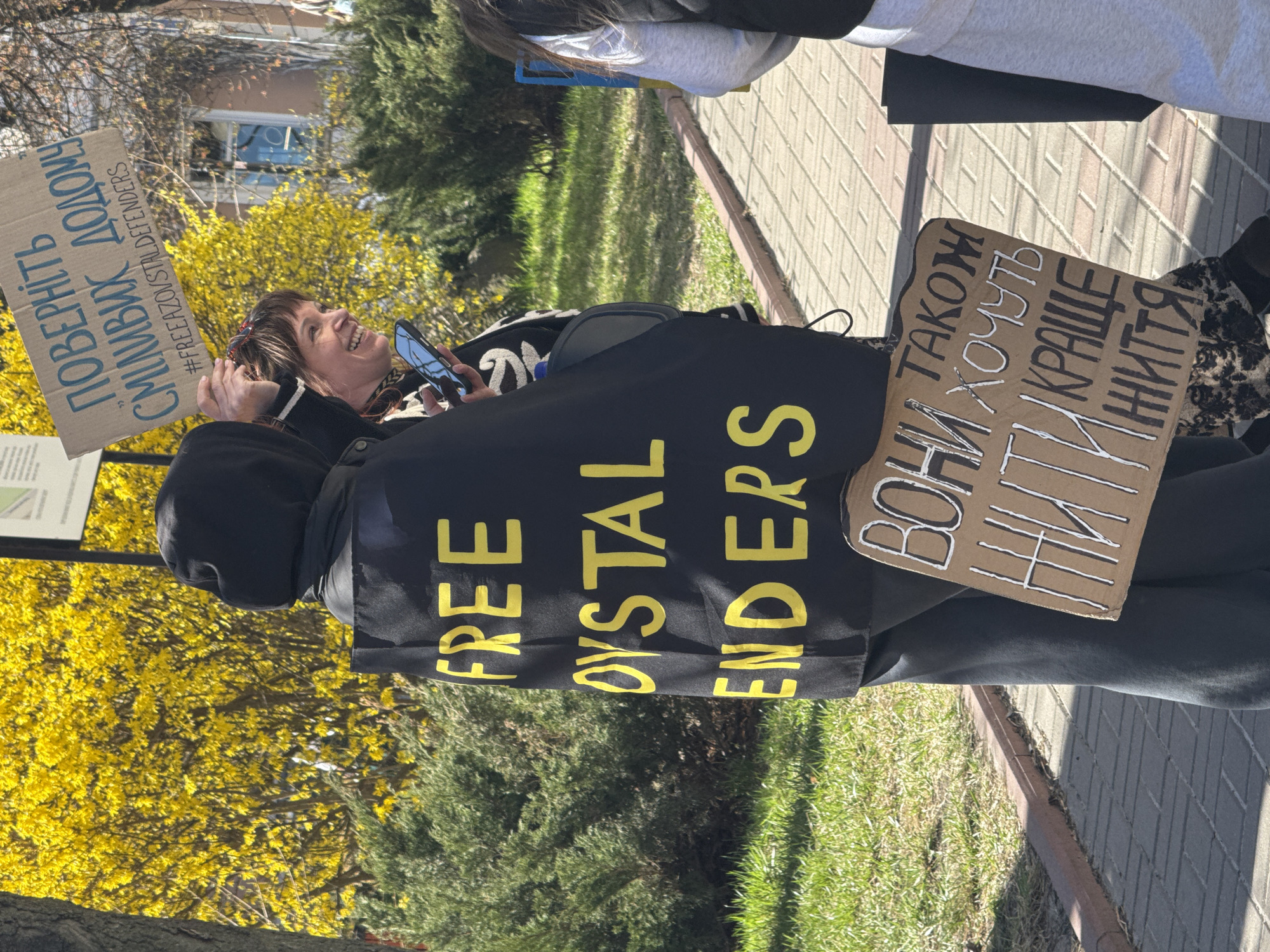 A medium, eye-level outdoor shot depicts at least three people protesting on a sunny day, likely in a park-like area within Kyiv, Ukraine, based on the presence of Ukrainian script on signage.
A woman in the center, facing left and looking up, holds a smartphone in her right hand and a cardboard sign in her left. The sign reads "ПОBEРНITЬ CMIЛИВИХ ДОДОМУ" (Return the brave home) and "#FREEAZOVSTALDEFENDERS." The woman has short brown hair and is smiling.
Behind her, an individual wears a black hooded sweatshirt and a black garment with large yellow lettering that reads "FREE OYSTAL ENDERS."
On the right side of the frame, another person holds a cardboard sign with Cyrillic script reading "ВОНИ ТАКОЖ ХОЧУТЬ КРАЩЕ ЖИТТЯ" (They also want a better life).
The background includes trees with bright yellow blossoms, a grassy area, and a paved walkway. A building with a window is visible in the upper left corner. The overall scene suggests a peaceful protest.
