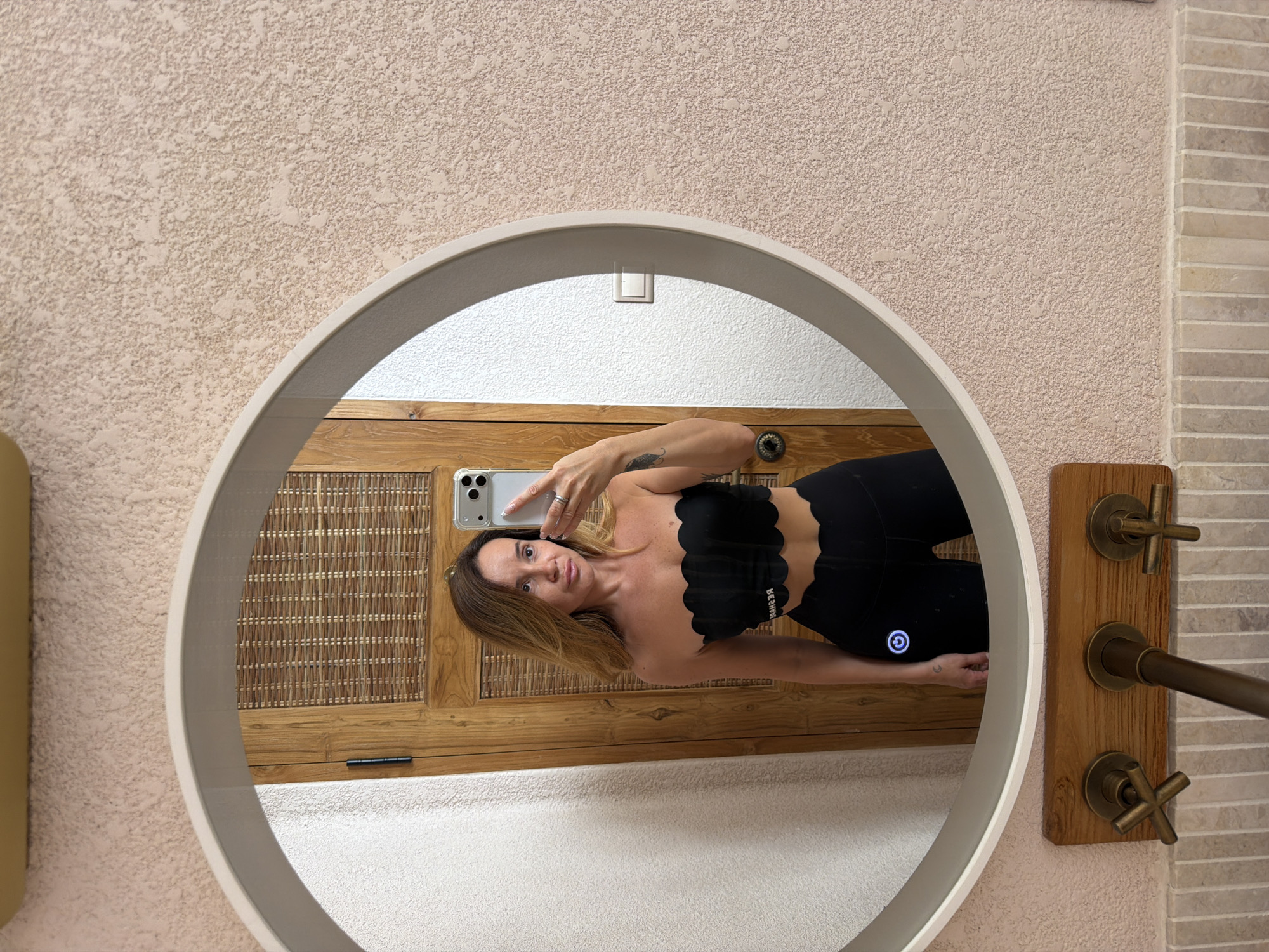 A mirror reflects a woman holding a phone, taking a selfie. She's wearing a black scalloped-edge strapless top and black leggings. The reflection also shows a wooden cabinet or door behind her and part of a tiled wall with wooden fixtures and antique-style brass faucets to the right of the mirror. The mirror has a white frame and the wall around it is textured. The image was taken in Mengwi, Indonesia.